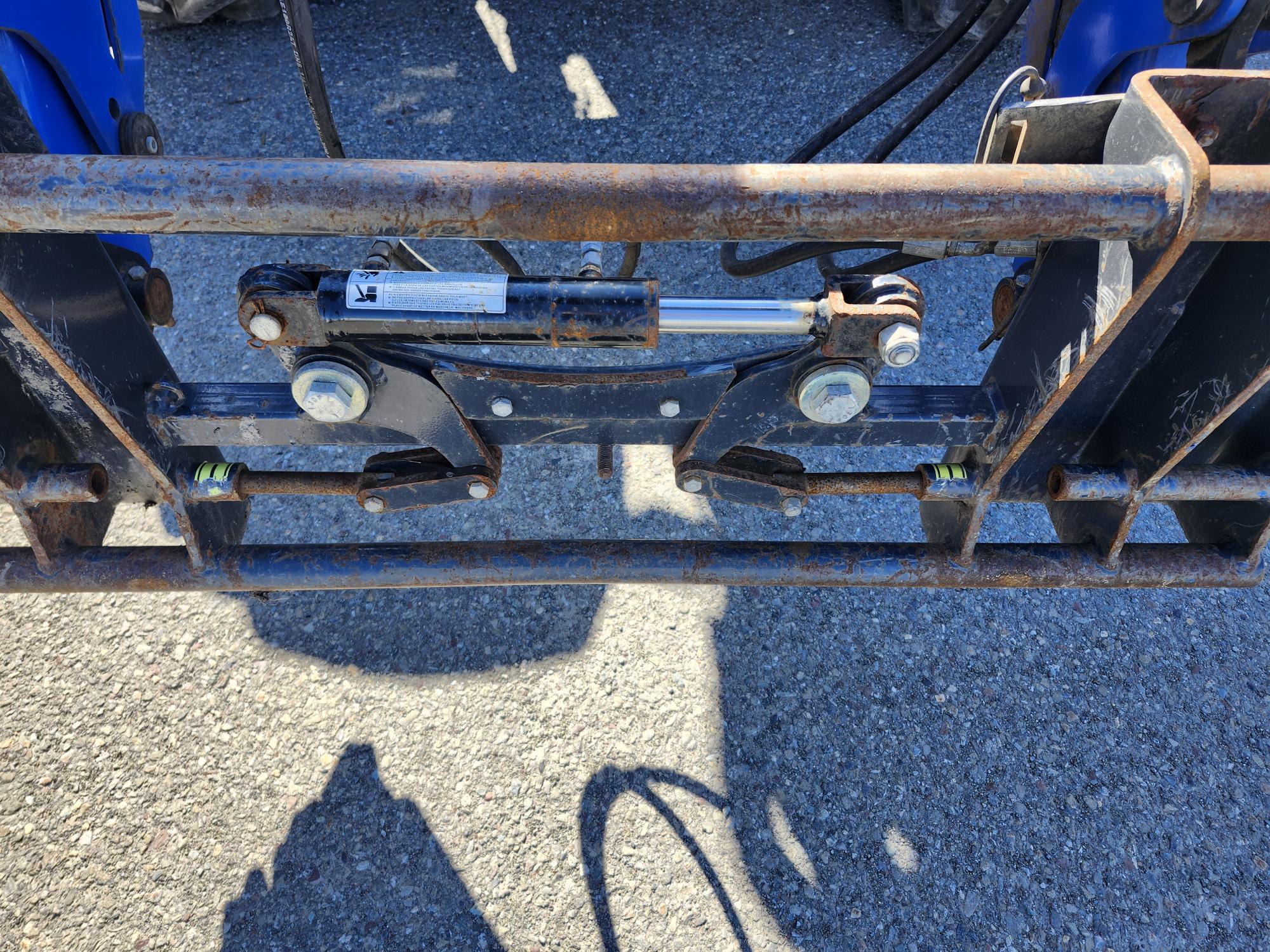 Gros plan d'un mécanisme hydraulique rouillé fixé à un cadre en métal bleu, reposant sur une surface asphaltée avec lumière du soleil et ombres visibles.