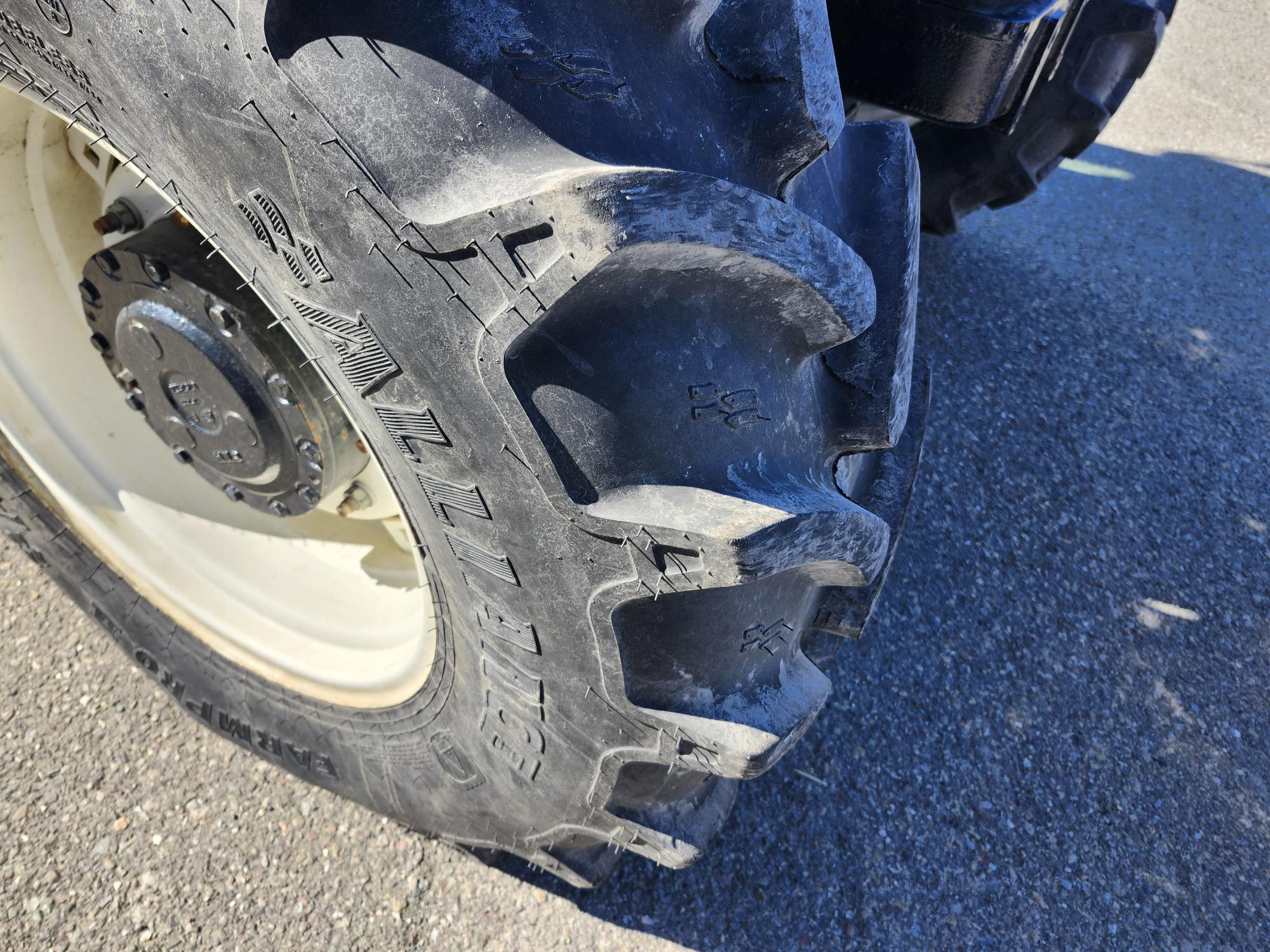 Vue rapprochée d'un gros pneu de tracteur robuste avec des bandes de roulement profondes sur une jante de roue noire, positionné sur une surface pavée en plein soleil.