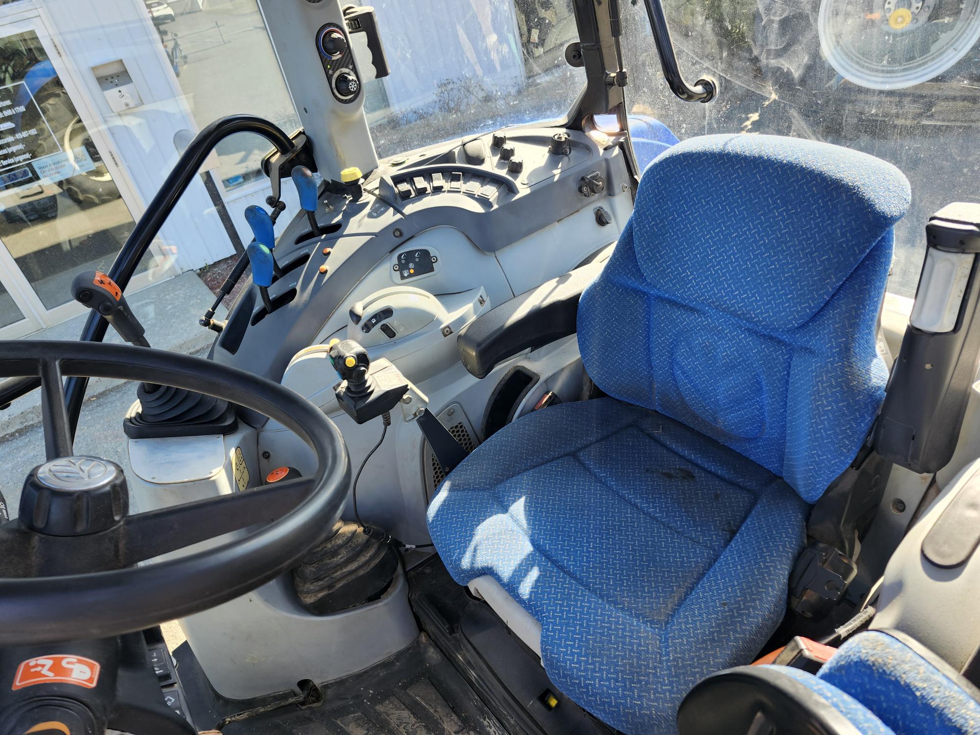 L'intérieur d'une cabine de tracteur, avec son siège conducteur en tissu bleu, son volant et les diverses commandes et leviers du tableau de bord. La lumière du soleil filtre à travers les vitres, illuminant l'aménagement moderne et épuré.