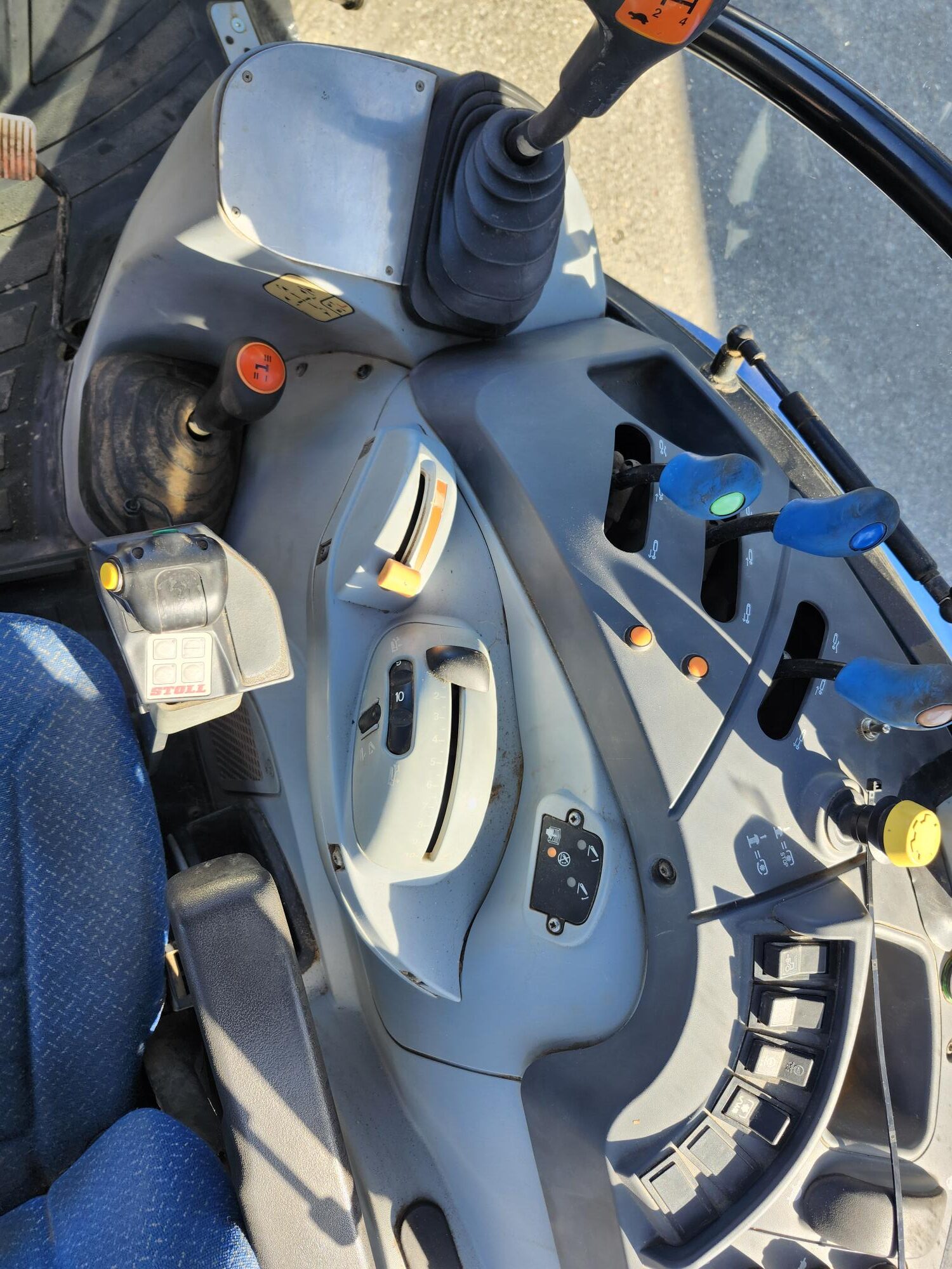 L'image montre le panneau de commande d'un tracteur, doté de multiples leviers, boutons, pédales et d'un volant. Le siège est recouvert d'un revêtement bleu à motifs et les commandes sont identifiées par divers symboles et couleurs.