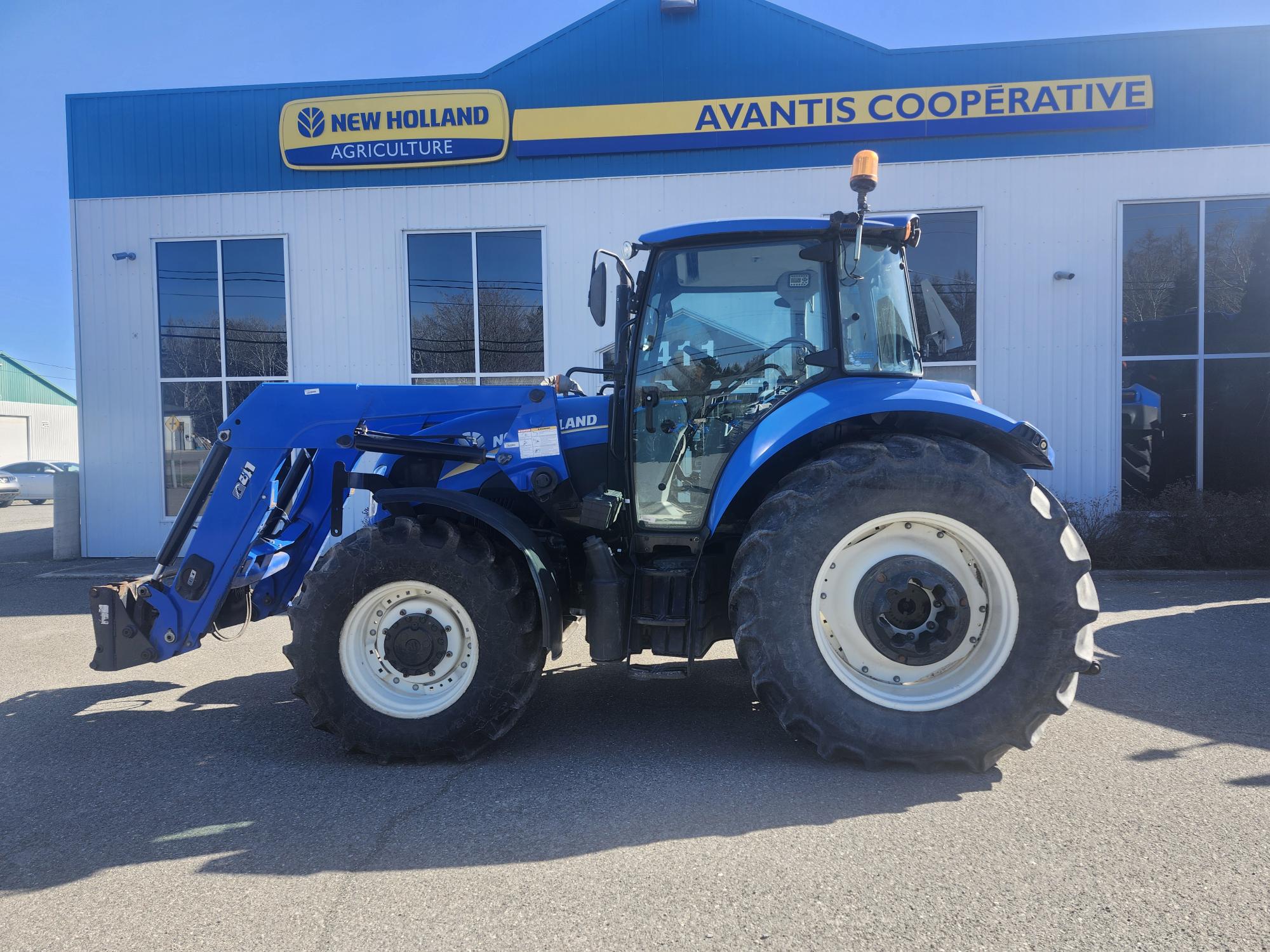 Un tracteur New Holland bleu est garé à l'extérieur d'un bâtiment avec de grandes fenêtres et un panneau indiquant « Avantis Cooperative » et « New Holland Agriculture ».
