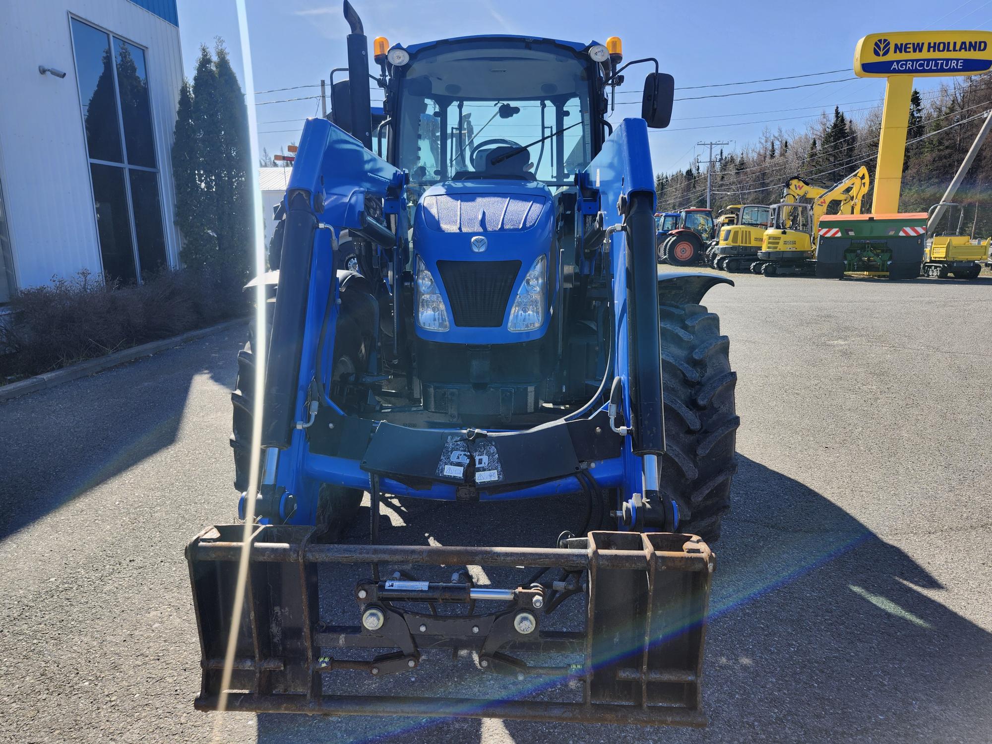 Un tracteur bleu équipé d'un chargeur frontal est garé à l'extérieur sur le trottoir près d'un concessionnaire New Holland Agriculture, avec d'autres machines visibles en arrière-plan sous un ciel clair.