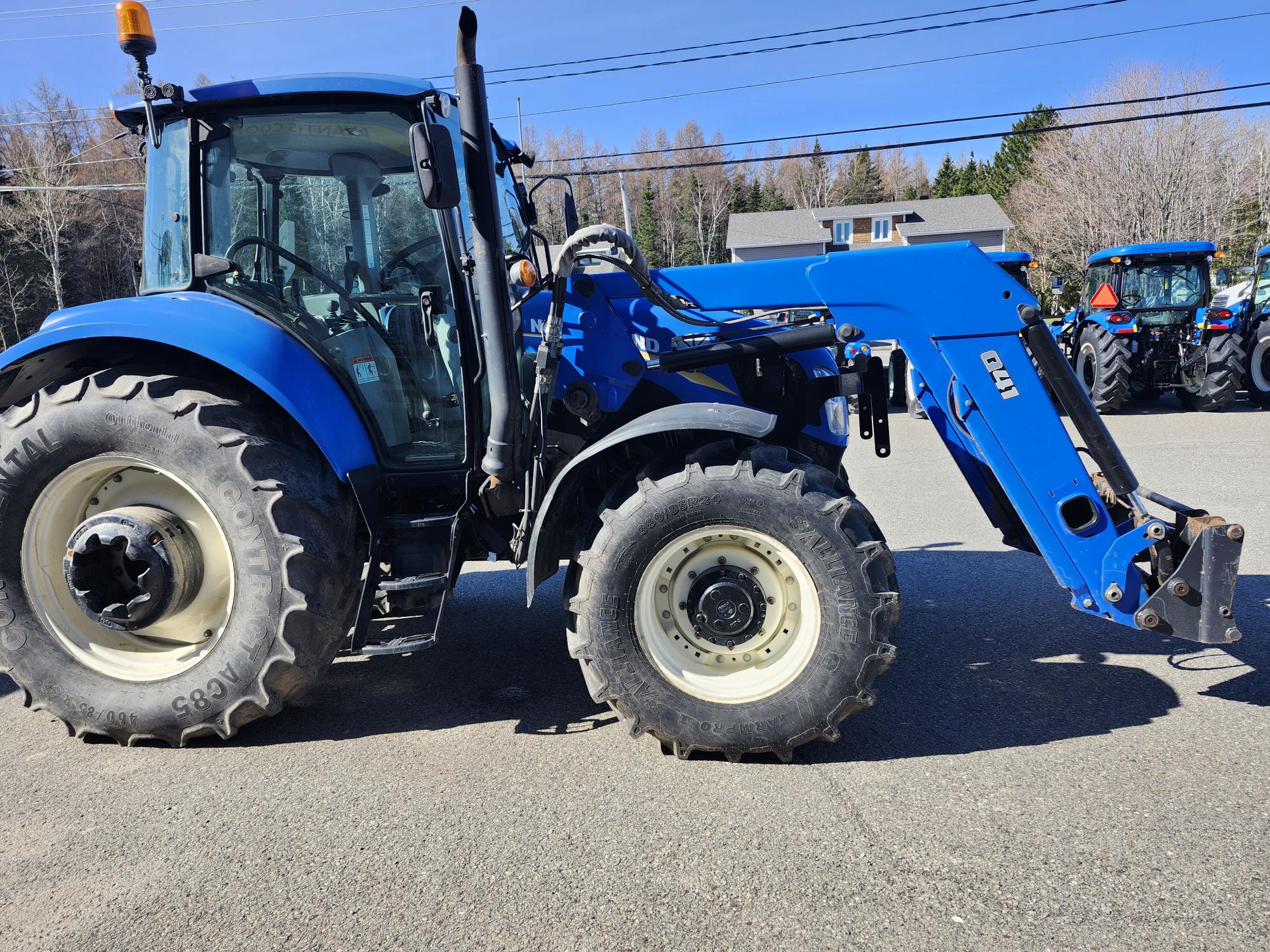 Un tracteur bleu avec de gros pneus et un chargeur frontal est garé sur le trottoir sous un ciel clair, avec des arbres et des bâtiments visibles en arrière-plan.