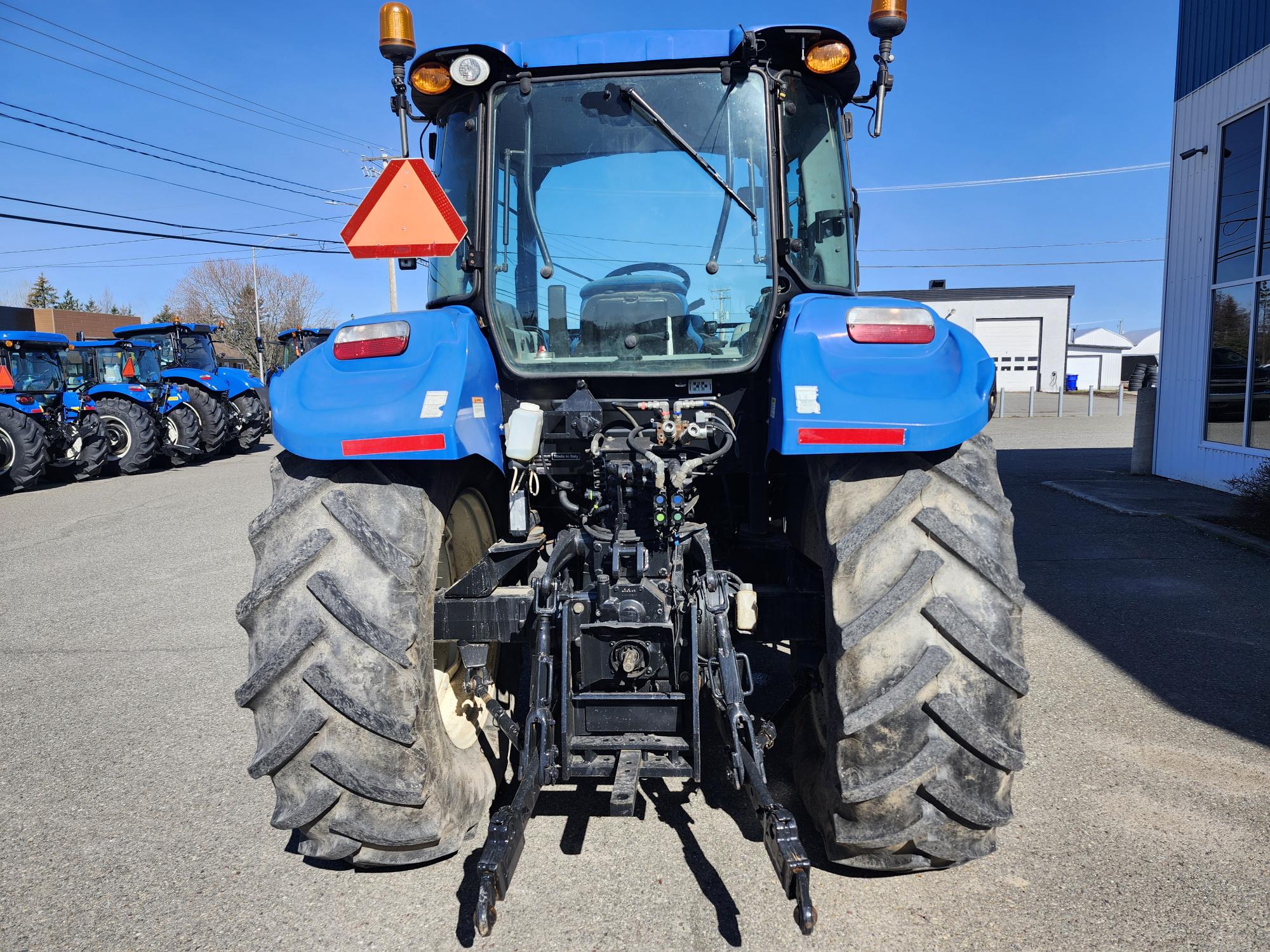 Vue arrière d'un tracteur bleu garé sur le trottoir près d'un bâtiment, montrant de gros pneus boueux, un panneau triangulaire réfléchissant rouge et divers composants mécaniques. D'autres tracteurs sont visibles en arrière-plan.