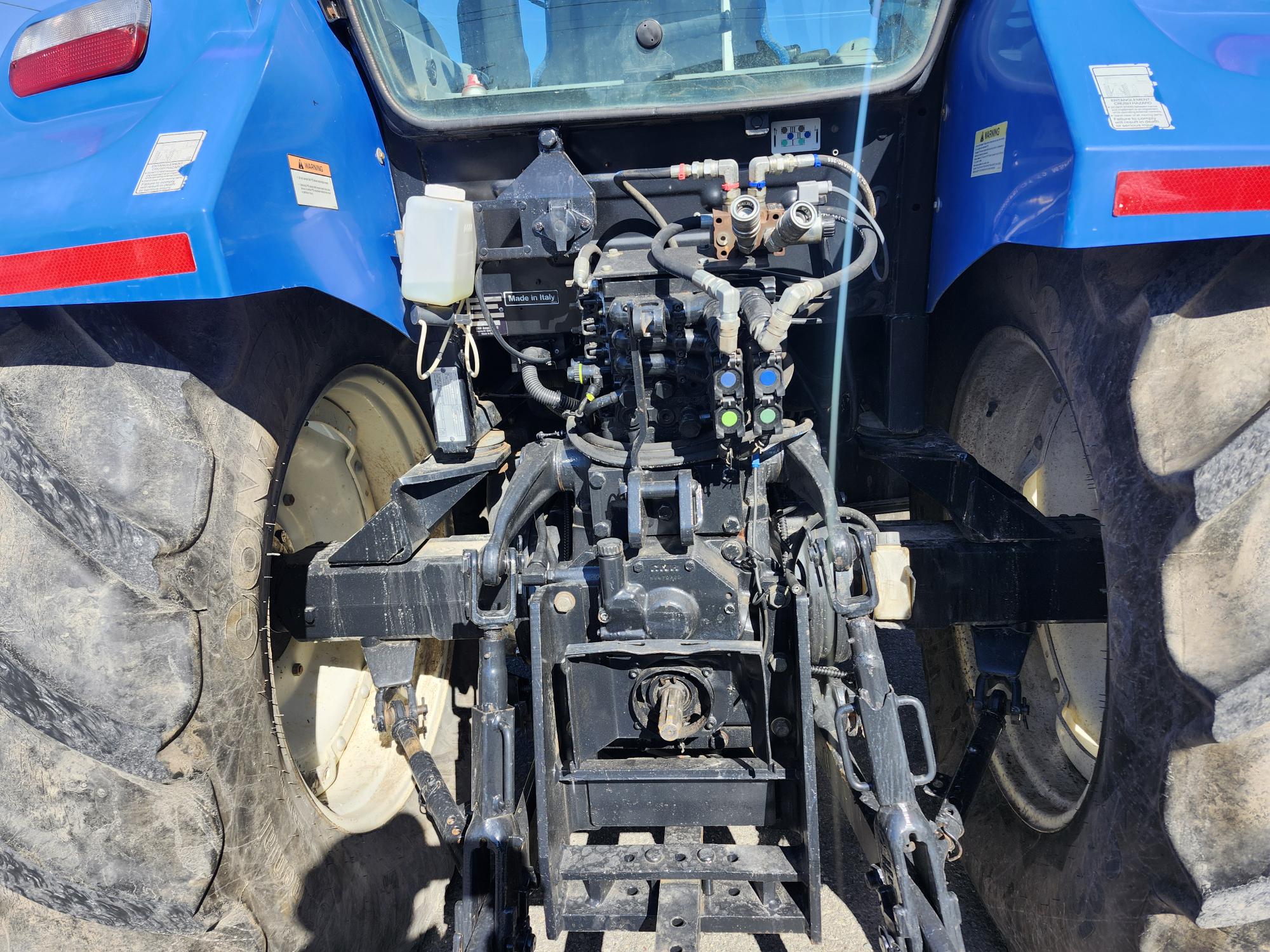 Vue rapprochée de l'arrière d'un tracteur bleu, montrant de gros pneus, des connexions hydrauliques et un ensemble d'attelage sous la lumière du soleil.