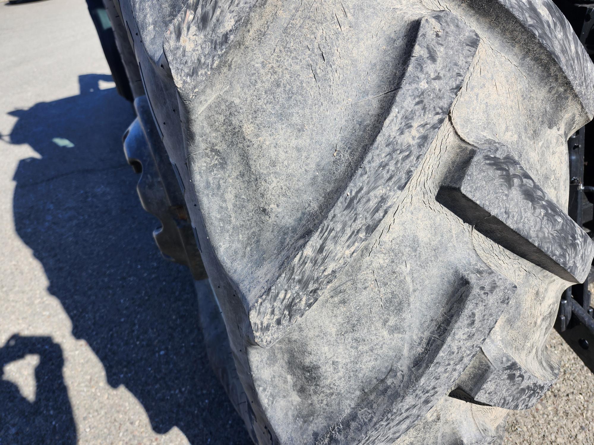 Gros plan d'un gros pneu de tracteur aux sculptures profondes et rugueuses. La surface du pneu présente des signes d'usure, et l'ombre d'une personne tenant une caméra est visible au sol à proximité.