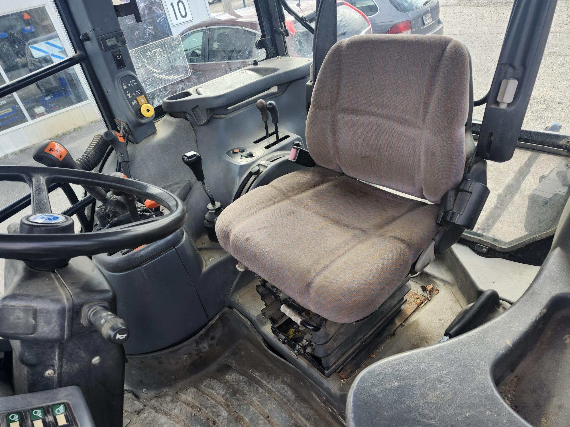 L'image montre l'intérieur d'une cabine de tracteur, avec un siège conducteur en tissu, un volant, divers leviers de commande et des pédales. Le sol paraît poussiéreux et les vitres laissent entrevoir des véhicules stationnés à l'extérieur.