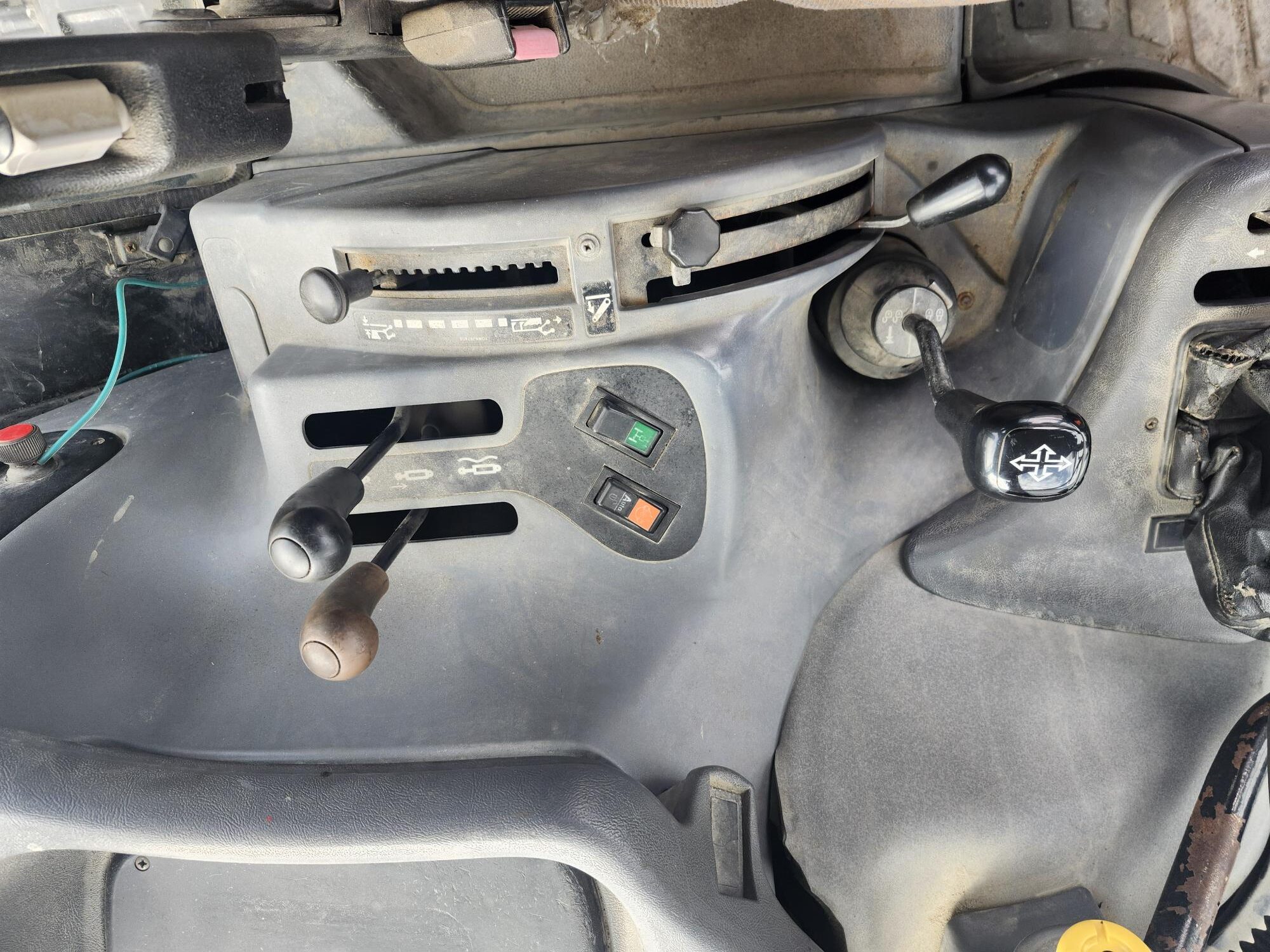 Vue du panneau de commande d'un tracteur ou d'une machine lourde, comportant divers leviers, interrupteurs et un levier de vitesses avec un motif d'engrenages sur le dessus. Le panneau est poussiéreux et présente des commandes mécaniques et des interrupteurs indicateurs.