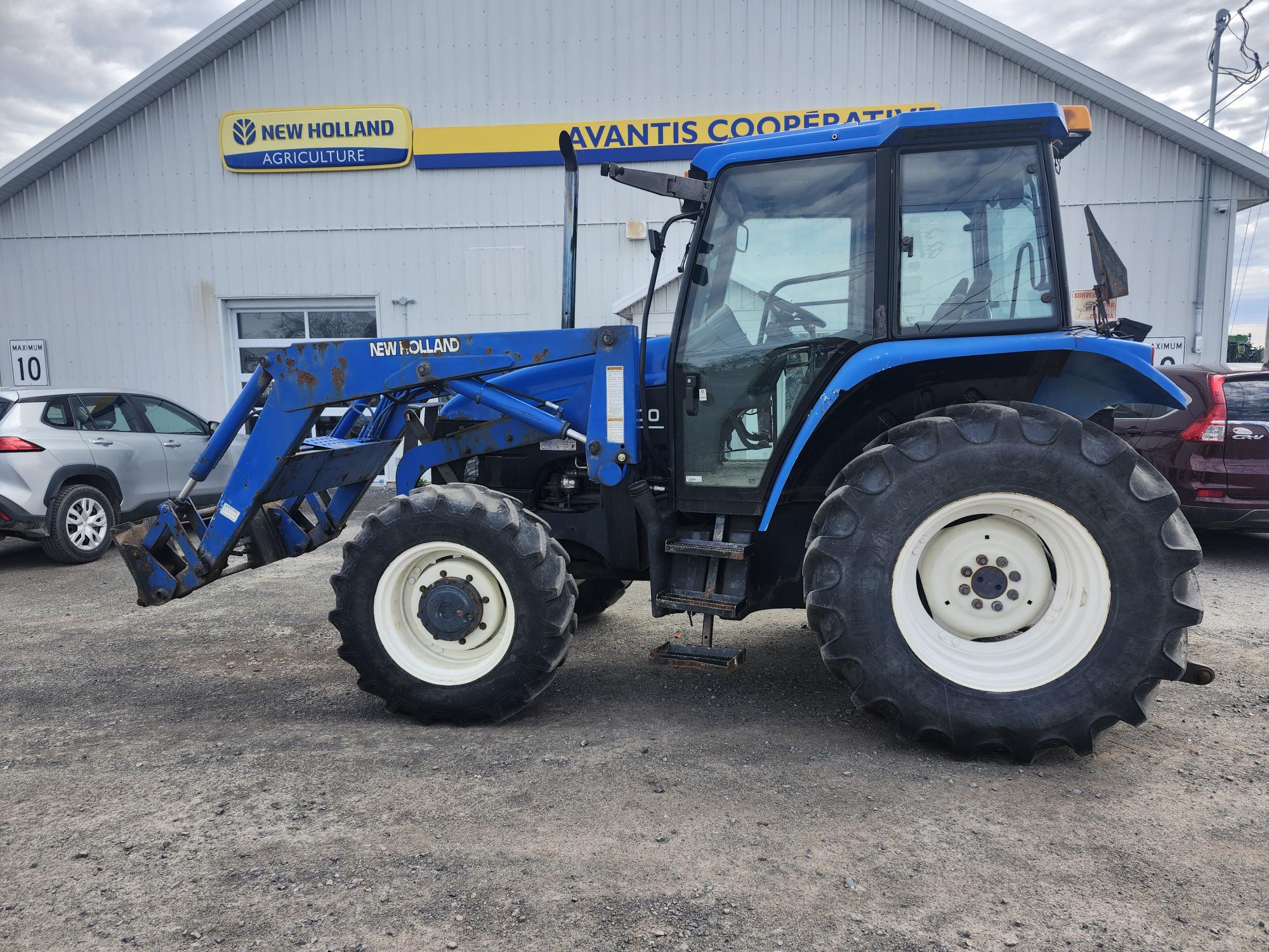 Un tracteur New Holland bleu équipé d'un chargeur frontal est garé sur du gravier à l'extérieur d'un bâtiment étiqueté « AVANTIS COOPERATIVE » et « NEW HOLLAND AGRICULTURE », avec plusieurs voitures en arrière-plan.