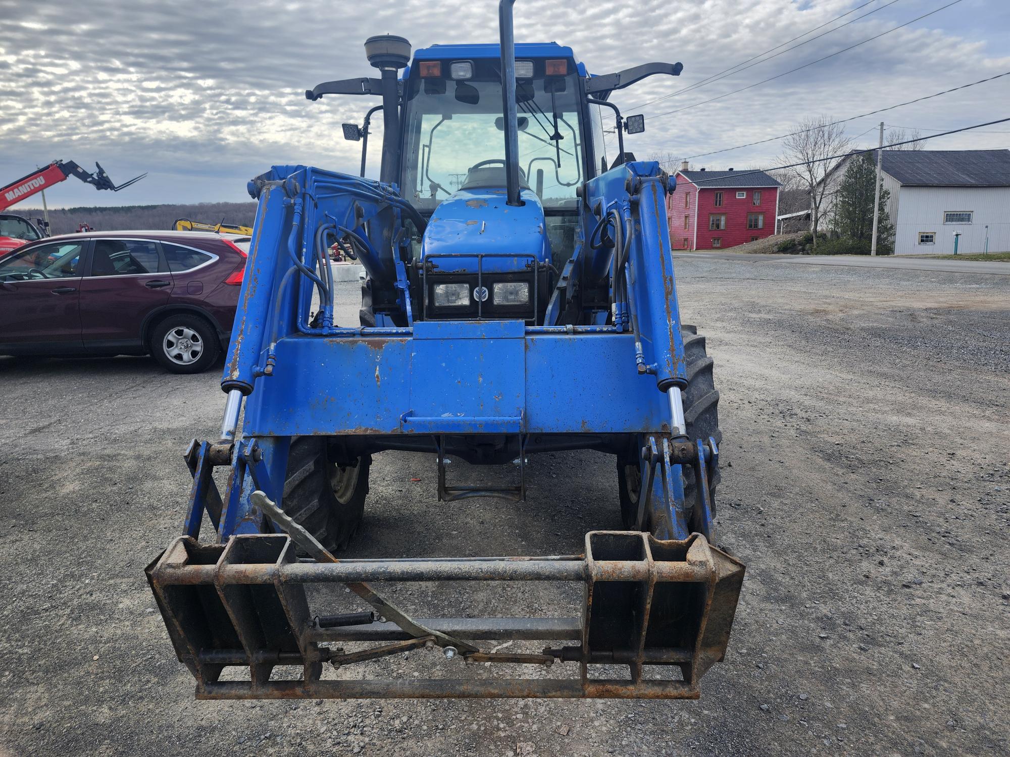 Un tracteur bleu équipé d'un chargeur frontal est garé sur un terrain en gravier, face à la caméra. À l'arrière-plan, plusieurs bâtiments, une voiture rouge et un ciel couvert.