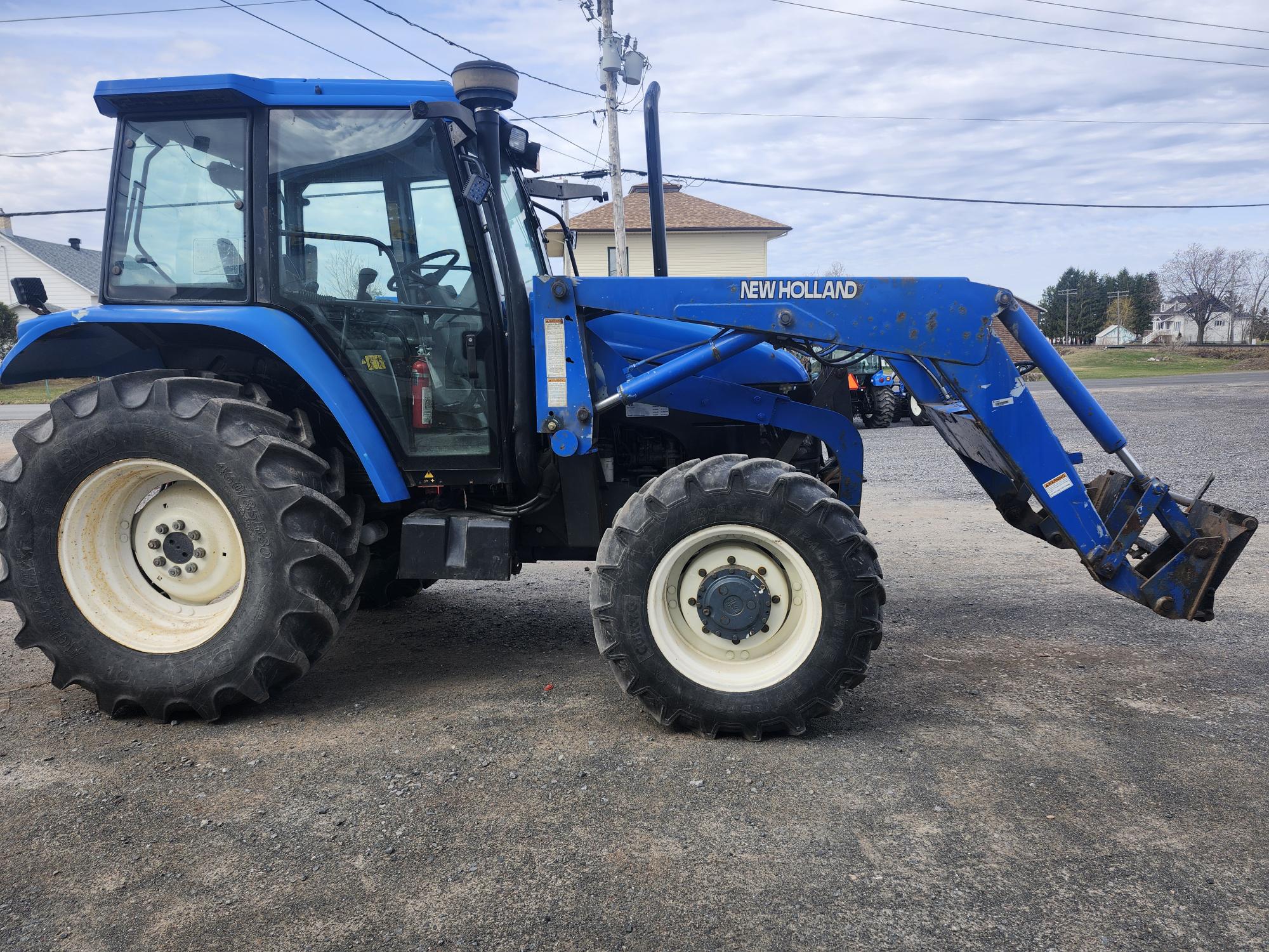 Un tracteur New Holland bleu équipé d'un chargeur frontal est garé sur une surface de gravier près de bâtiments et de lignes électriques, sous un ciel partiellement nuageux.
