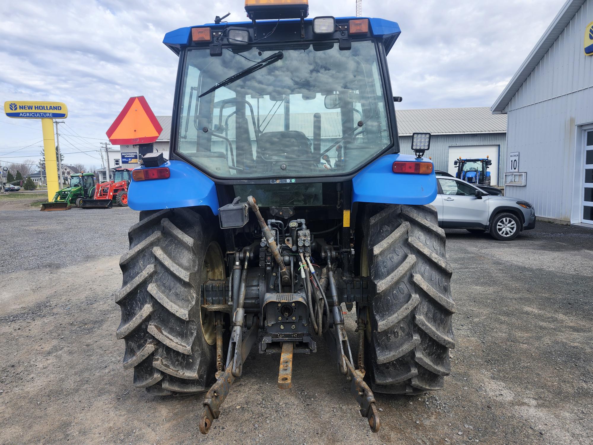 Un tracteur bleu vu de derrière, garé sur du gravier près d'un bâtiment blanc et d'un panneau New Holland Agriculture, avec un autre tracteur et une voiture grise visibles en arrière-plan.