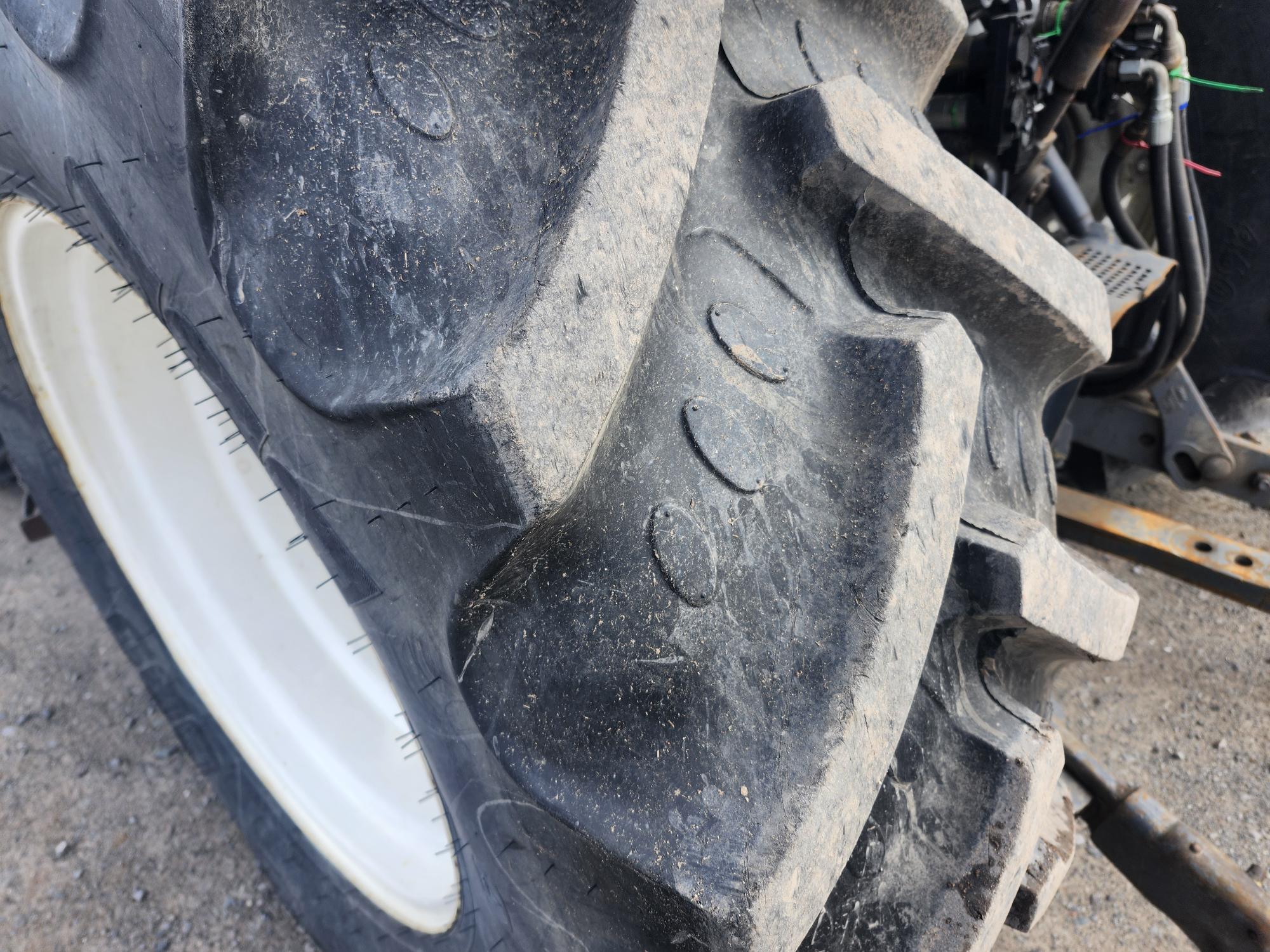 Gros plan sur un gros pneu de tracteur noir aux sculptures profondes, laissant apparaître des traces de saleté et d'usure. Une partie du châssis du tracteur et du sol gravillonné sont visibles en arrière-plan.