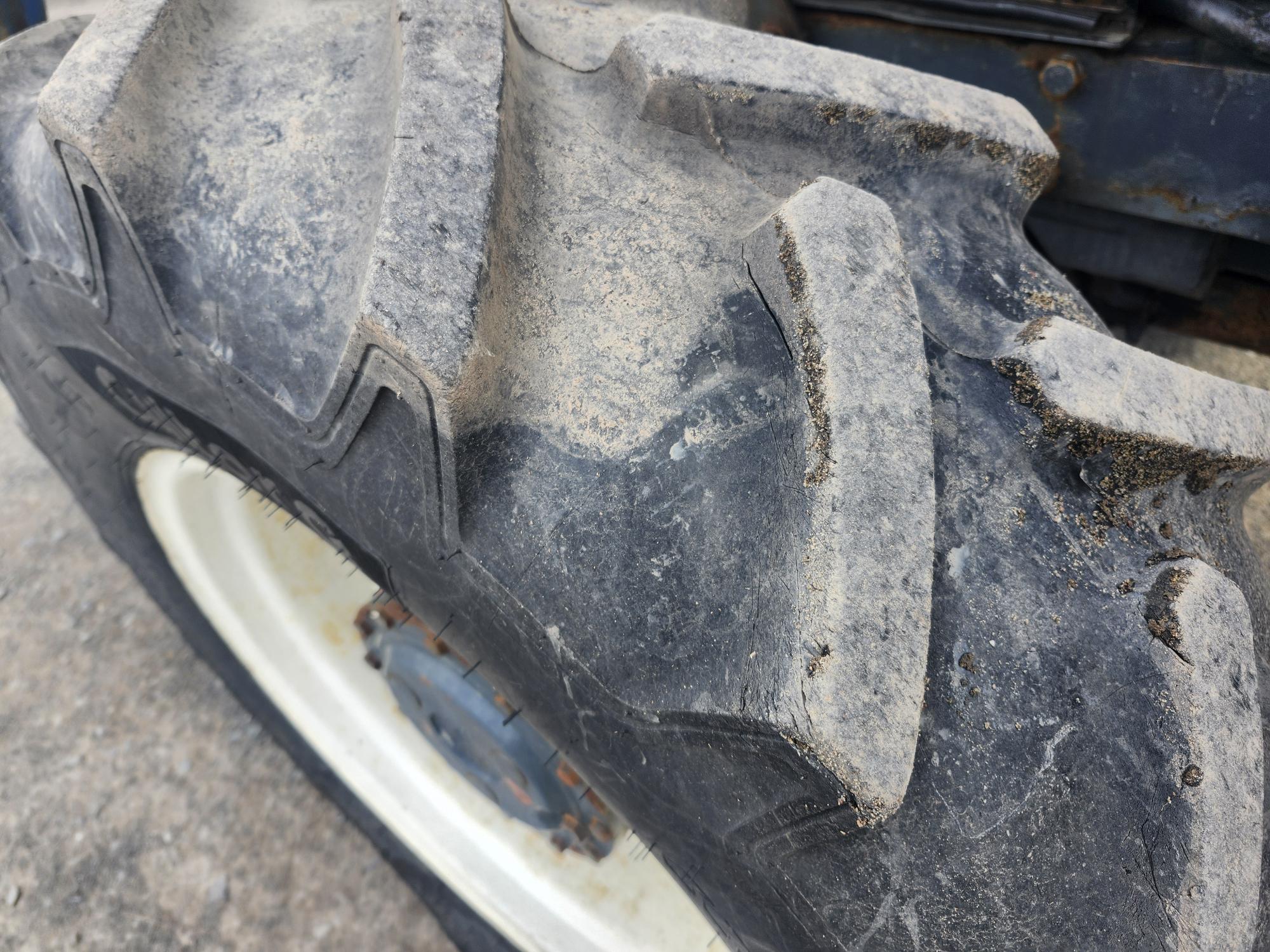 Gros plan d'un gros pneu de tracteur poussiéreux aux sculptures profondes, monté sur une jante blanche. Le sol en dessous est rugueux et gris, et le pneu présente des signes d'usure et de saleté.