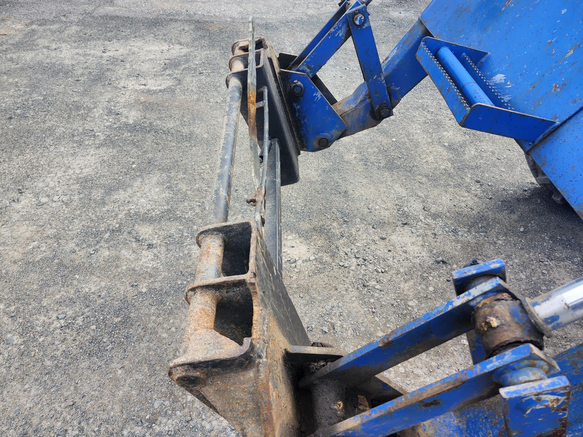 Gros plan d'un accessoire métallique rouillé sur un engin de chantier bleu, probablement une chargeuse ou un tracteur, posé sur une surface rugueuse en gravier. L'accessoire présente des signes d'usure et de corrosion.