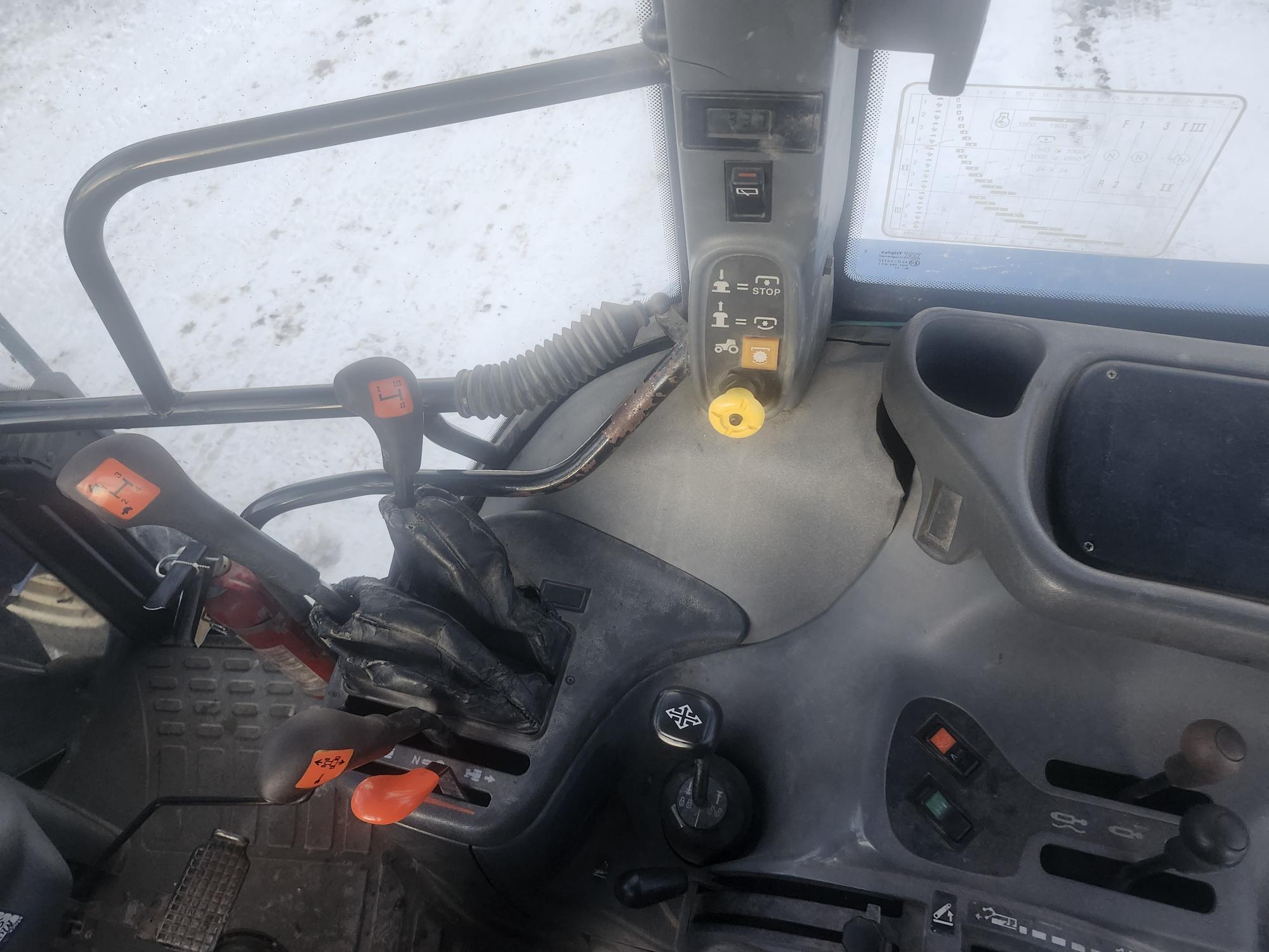 Vue intérieure d'une cabine de tracteur montrant diverses commandes, des leviers à poignées rouges et un bouton jaune. Des gants noirs reposent sur un levier. Un câble spiralé pend près d'un panneau de commande. De la neige est visible par la fenêtre.
