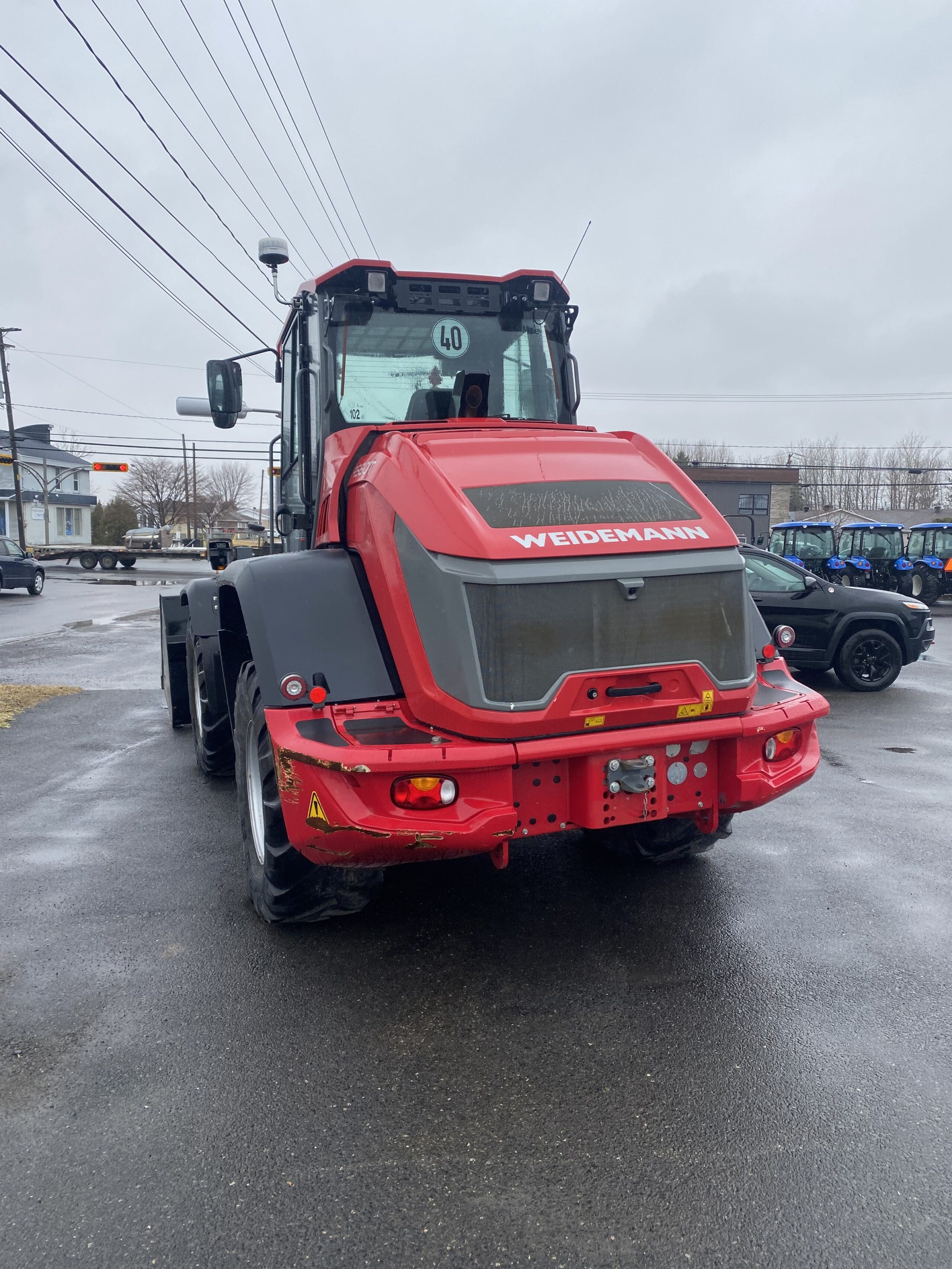 Un gros tracteur Weidemann rouge est garé sur un trottoir mouillé par temps nuageux, avec quelques bâtiments, lignes électriques et véhicules visibles en arrière-plan.