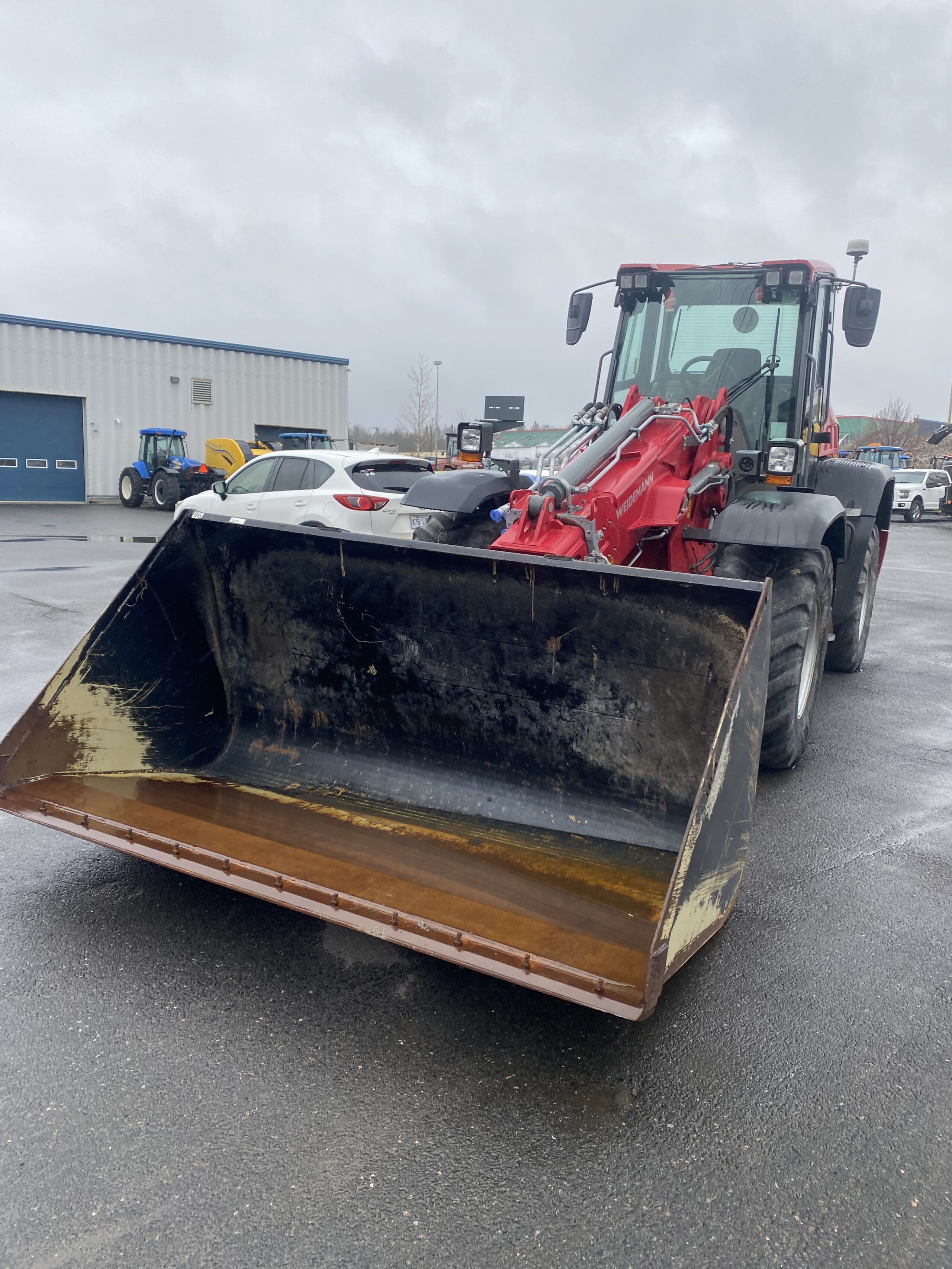 Un gros tracteur à chargeur frontal rouge et noir est garé sur la chaussée mouillée d'un parking, la benne relevée. Plusieurs voitures et un bâtiment industriel sont visibles en arrière-plan sous un ciel nuageux.