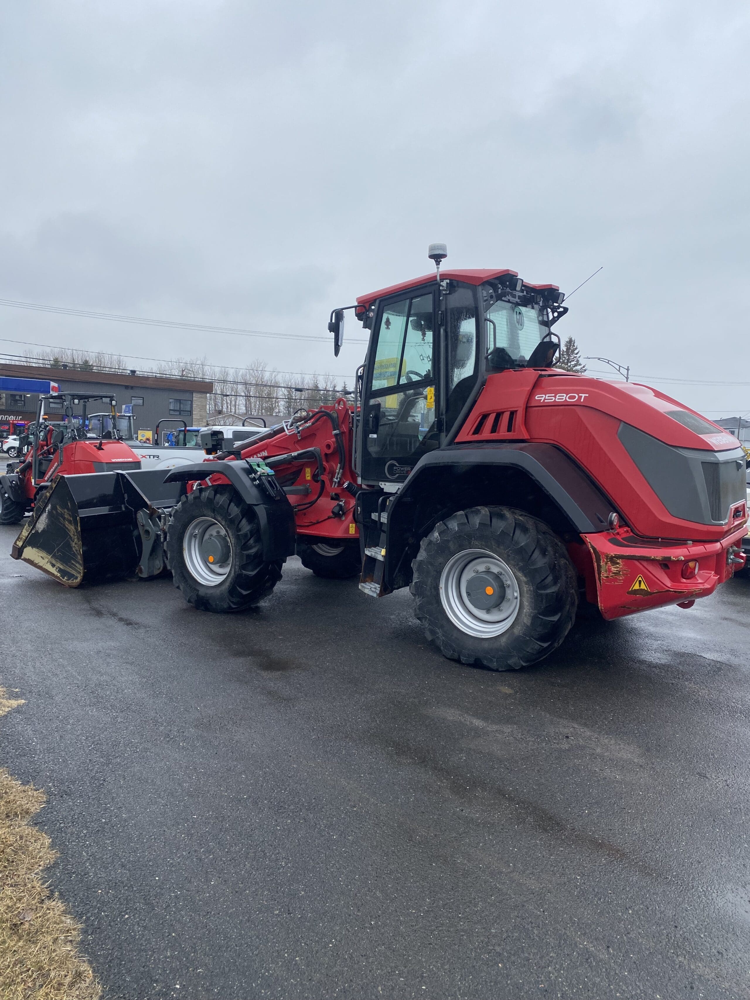 Un tracteur rouge Massey Ferguson 9560T équipé d'un chargeur frontal est stationné sur une surface asphaltée mouillée par temps nuageux. D'autres machines et bâtiments sont visibles en arrière-plan.