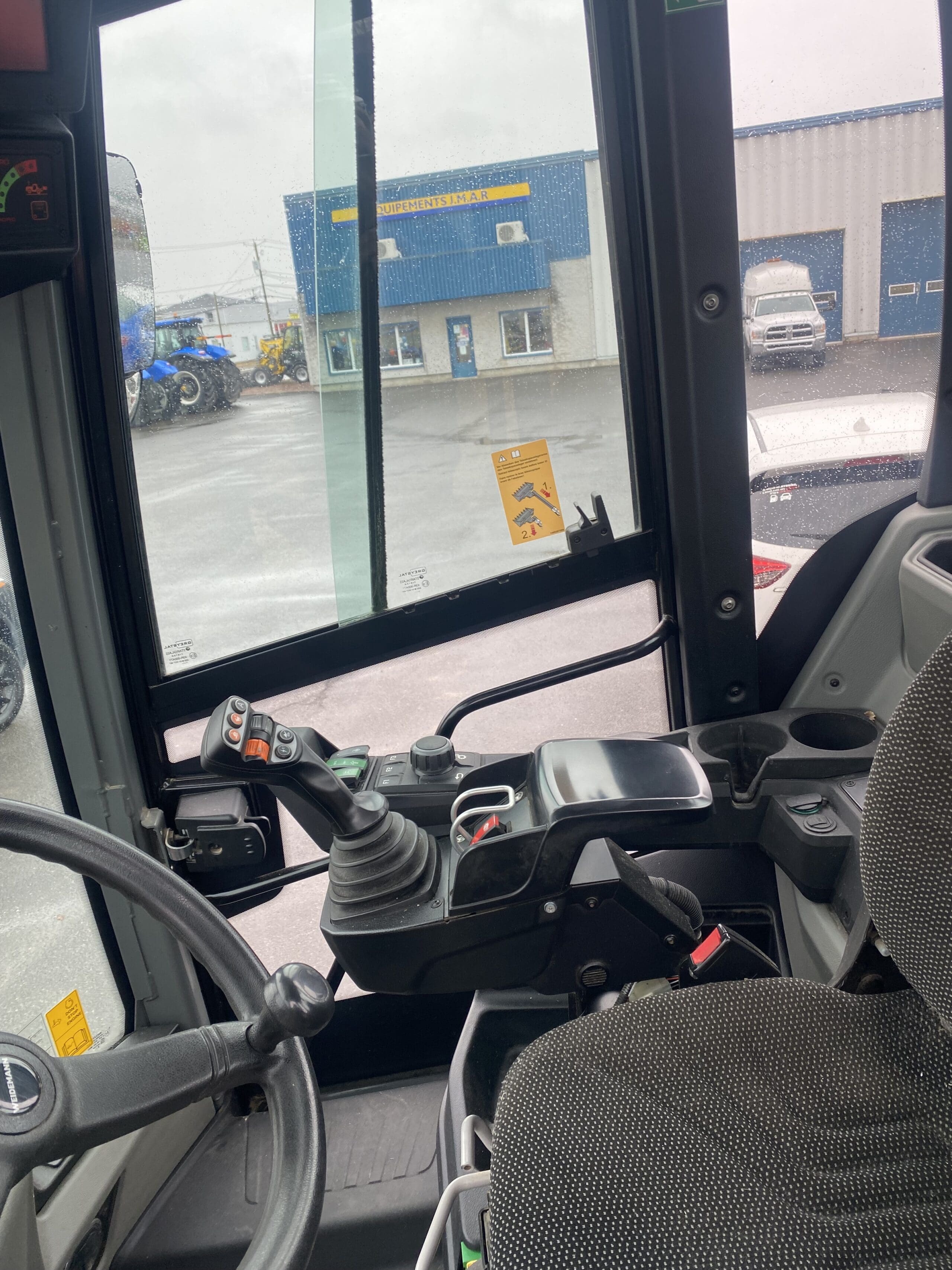 Vue de l'intérieur de la cabine d'un tracteur, montrant le volant, le panneau de commande, le joystick et le siège. À l'extérieur, on aperçoit un parking pluvieux et un bâtiment bleu à travers de grandes fenêtres.