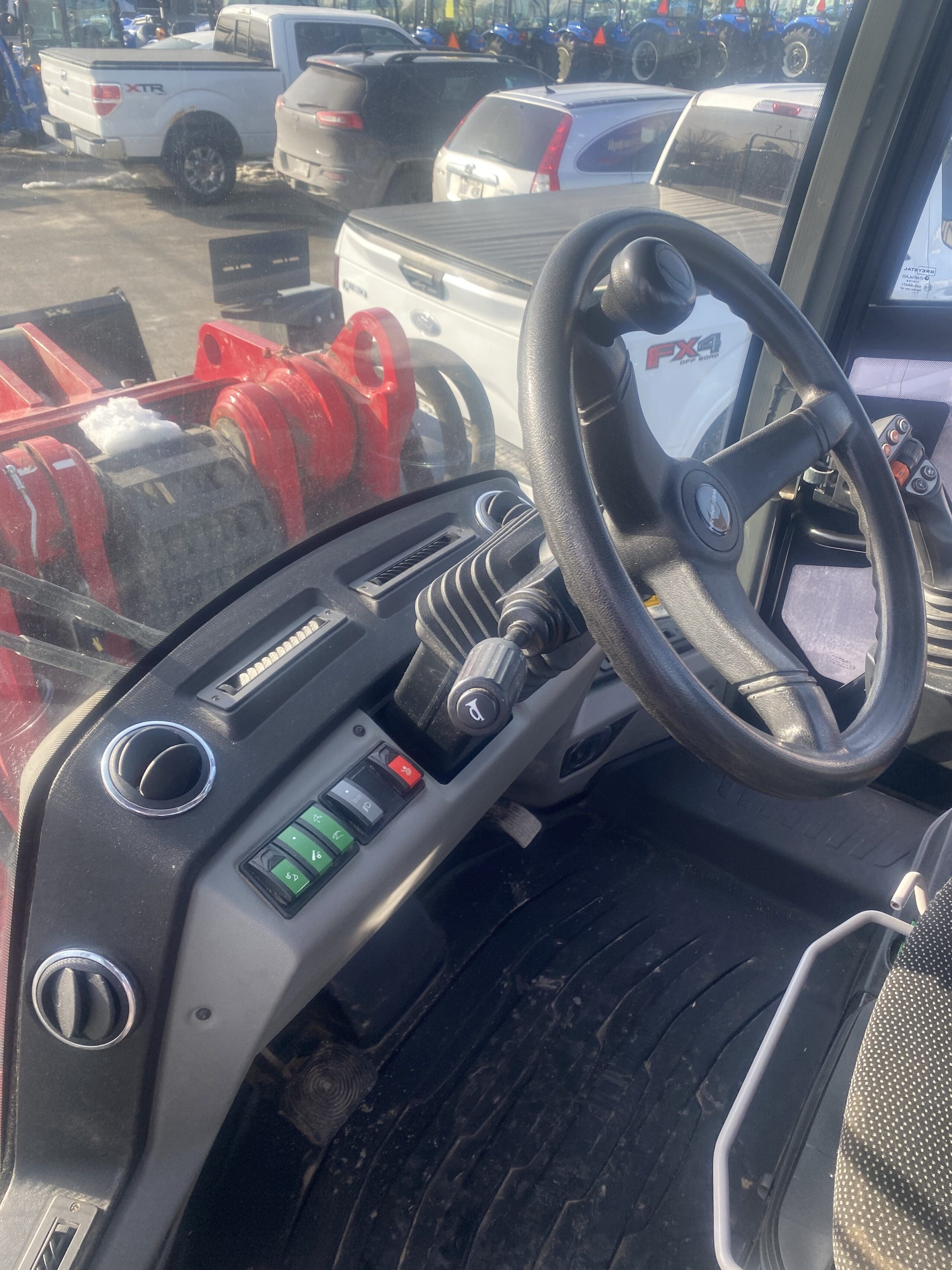 Vue rapprochée du siège du conducteur et du panneau de commande à l'intérieur d'un véhicule utilitaire, avec un volant, un levier de vitesses, des interrupteurs et des tapis de sol boueux ; plusieurs véhicules stationnés sont visibles à l'extérieur de la fenêtre.