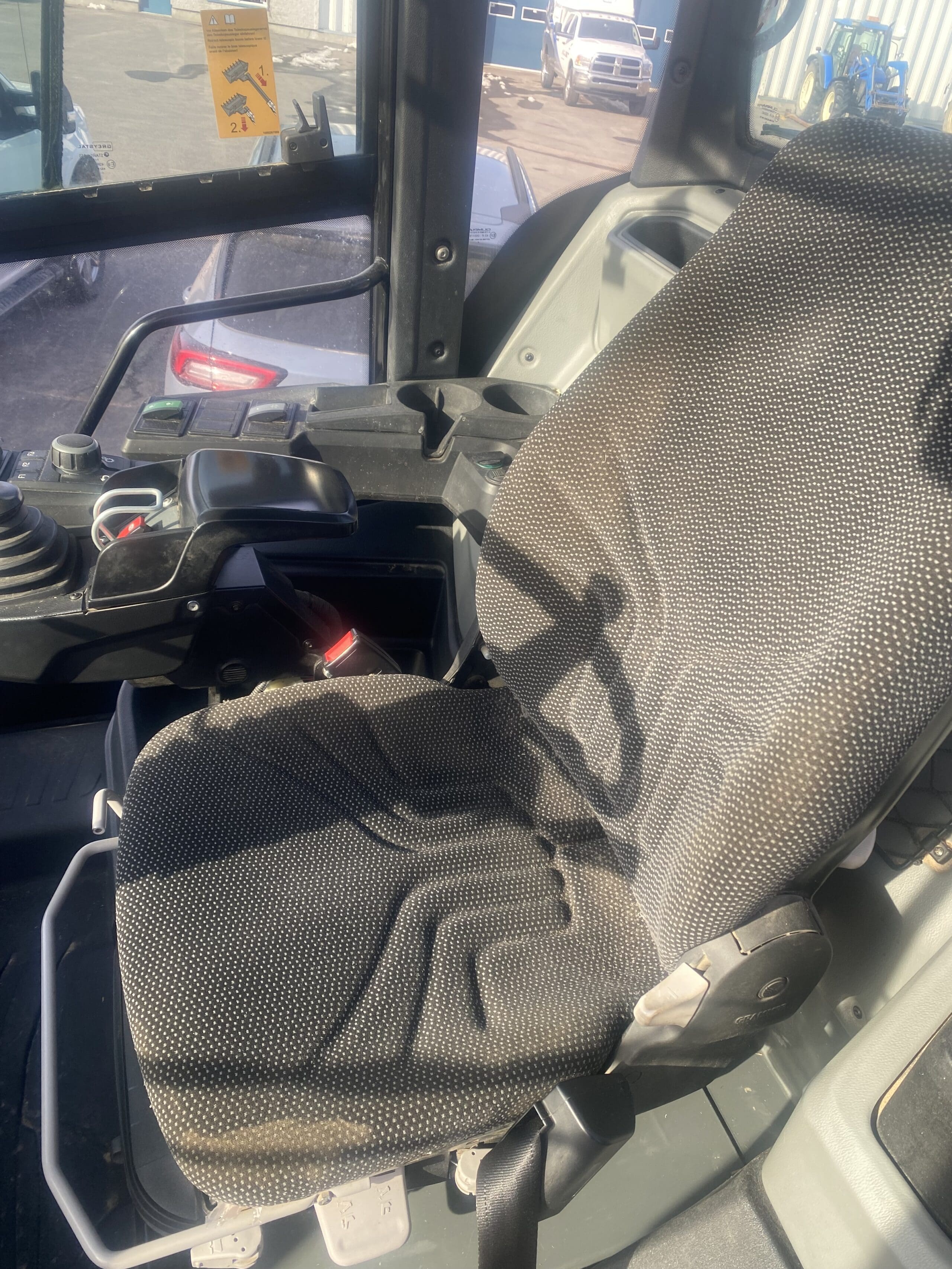 Siège conducteur et panneau de commande à l'intérieur d'une cabine d'engin de chantier, avec sellerie grise texturée et leviers de commande visibles. La lumière du soleil filtre à travers les vitres, mettant en valeur le siège et les commandes.