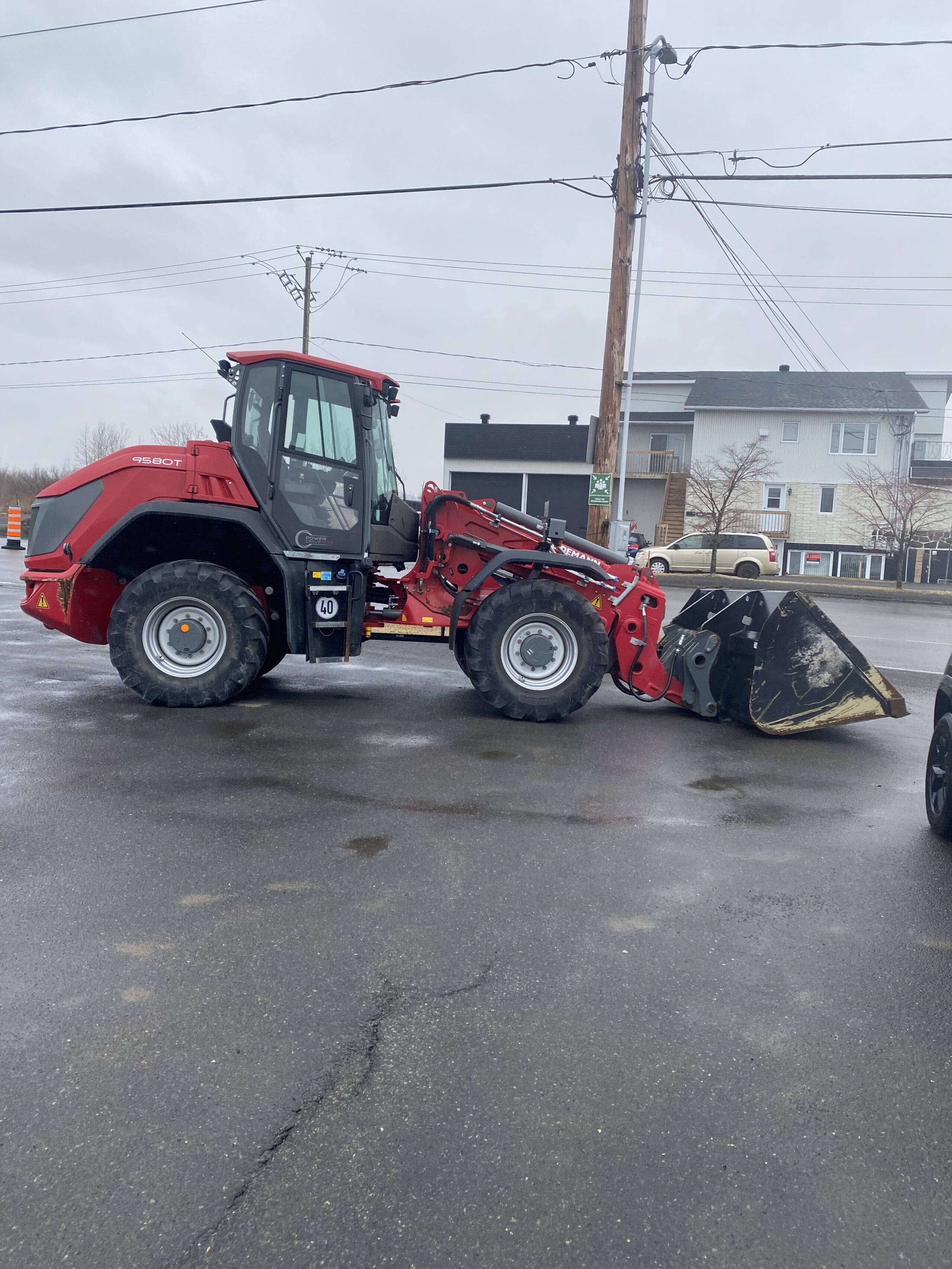 Un engin de chantier rouge à chargeur frontal est garé dans une rue humide près d'un quartier résidentiel avec des maisons à plusieurs étages et plusieurs voitures garées en arrière-plan. Le ciel est couvert.
