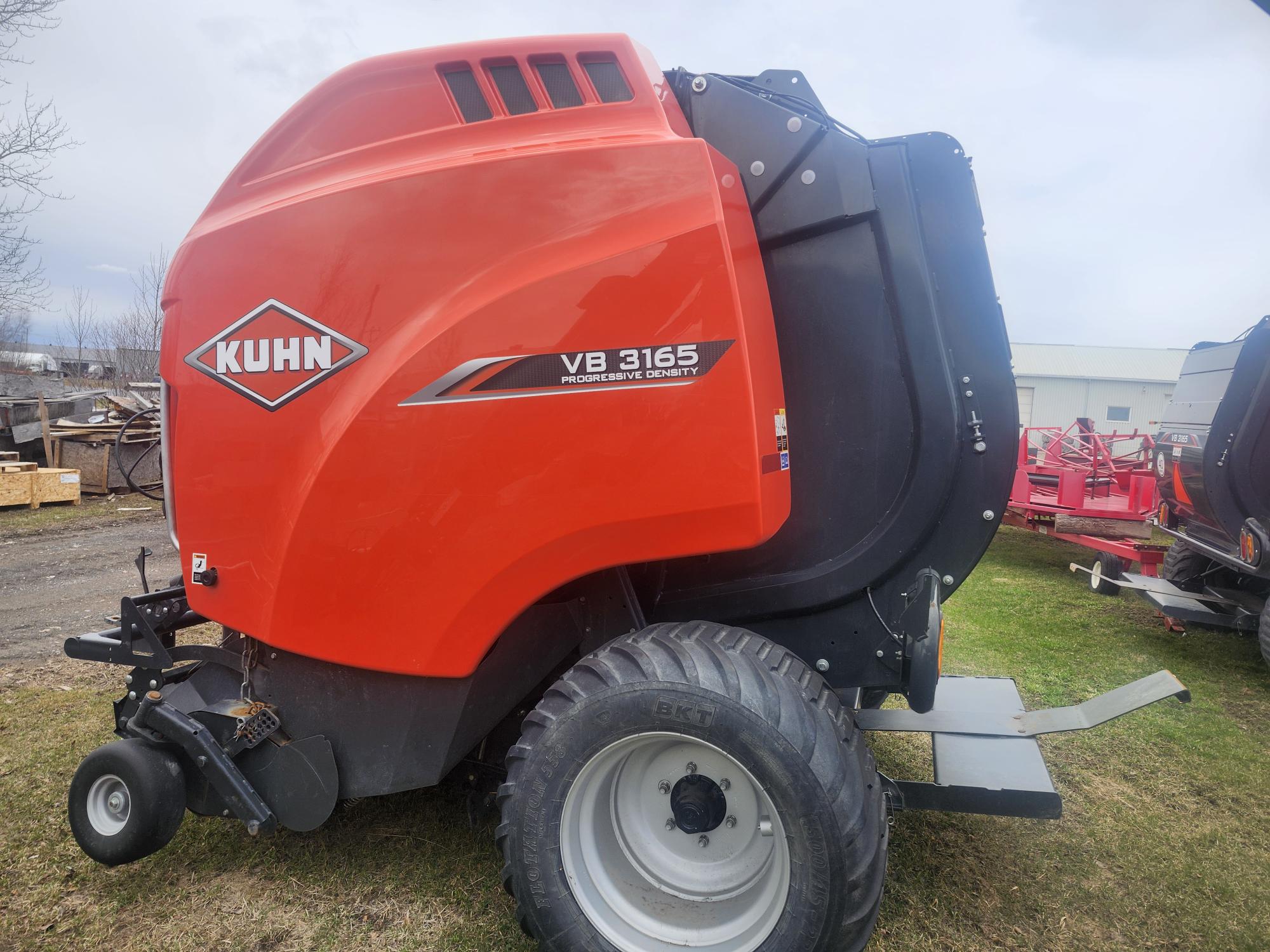 Une presse à balles rondes KUHN VB 3165 rouge est posée sur l'herbe, laissant apparaître sa grande roue, ses mécanismes métalliques et son logo sur le côté. Des équipements agricoles et des bâtiments sont visibles en arrière-plan.