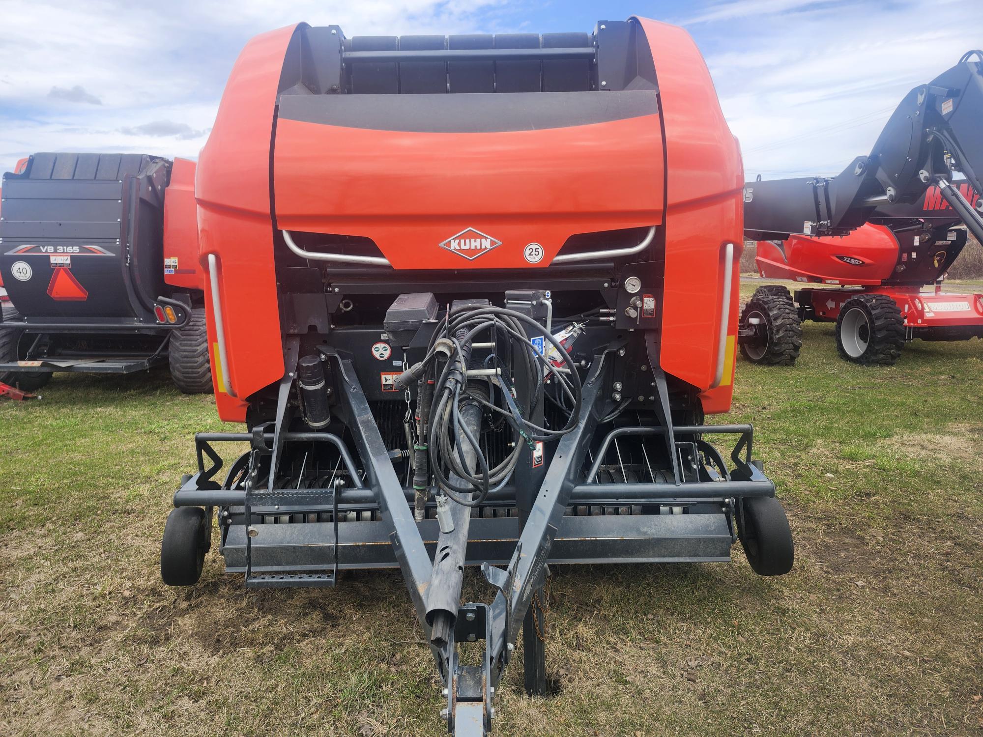 Une grande presse agricole Kuhn orange est exposée en extérieur sur l'herbe. La machine est équipée de diverses pièces métalliques et de flexibles hydrauliques, et est entourée d'autres équipements agricoles sous un ciel partiellement nuageux.