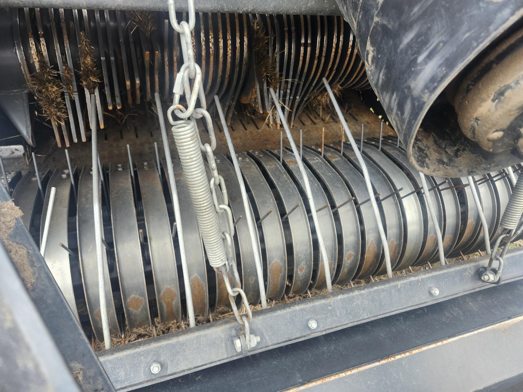 Vue rapprochée d'une pièce de machine mécanique comportant des disques métalliques avec des dents saillantes et pointues, un grand ressort et des chaînes ; faisant probablement partie d'un équipement agricole ou industriel.