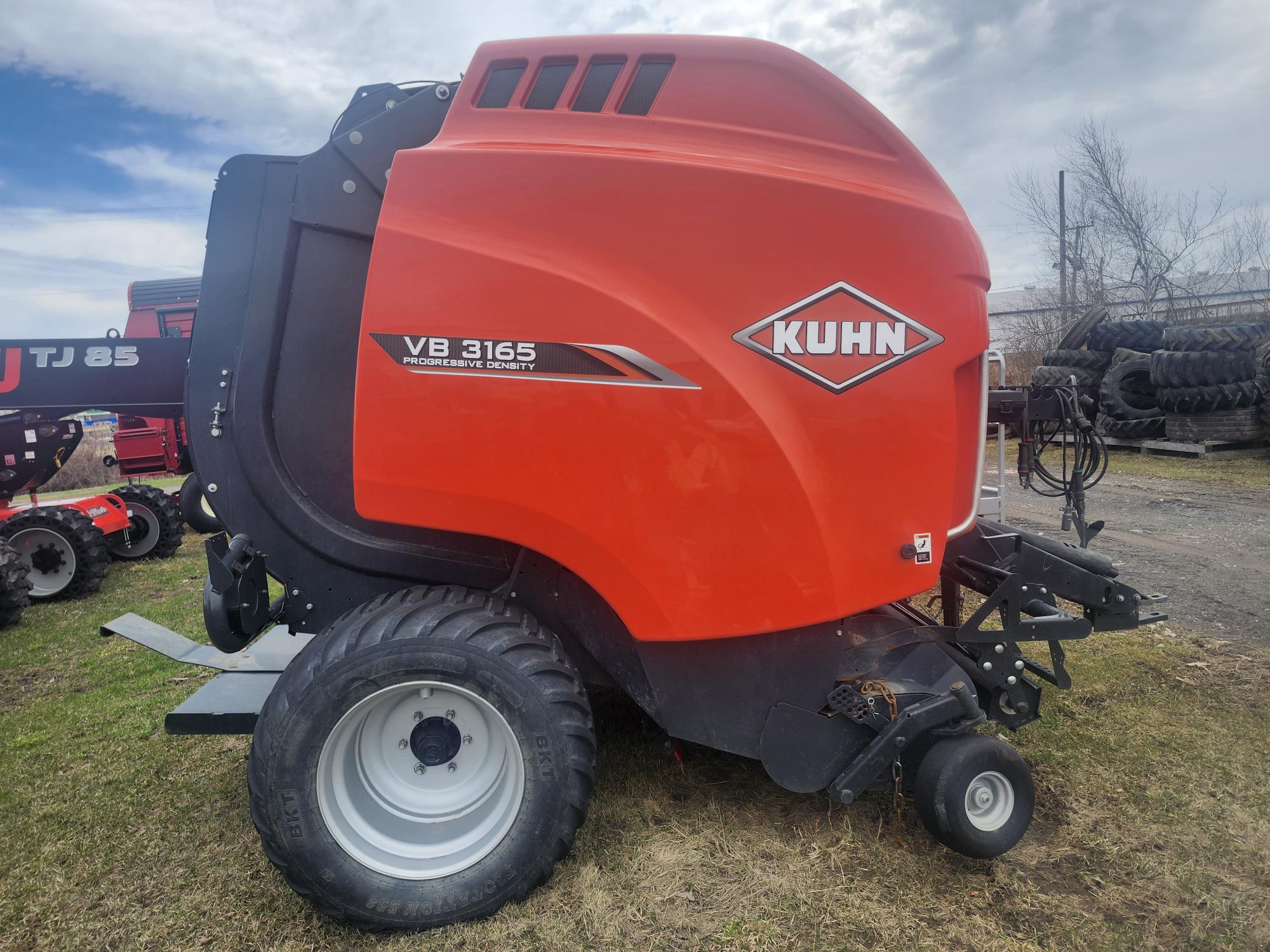 Une presse à balles rondes Kuhn VB 3165 orange vif repose sur l'herbe, avec de gros pneus noirs et un ciel nuageux en arrière-plan. La machine porte le logo Kuhn et d'autres machines sont partiellement visibles à proximité.