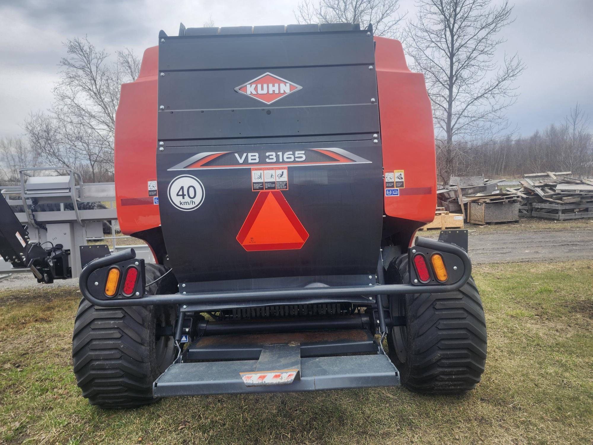 Une grande presse agricole Kuhn VB 3165 rouge et noire est garée sur l'herbe, avec des autocollants d'avertissement et un panneau de limitation de vitesse à 40 km/h à l'arrière. Des arbres dénudés et des palettes en bois sont visibles en arrière-plan.