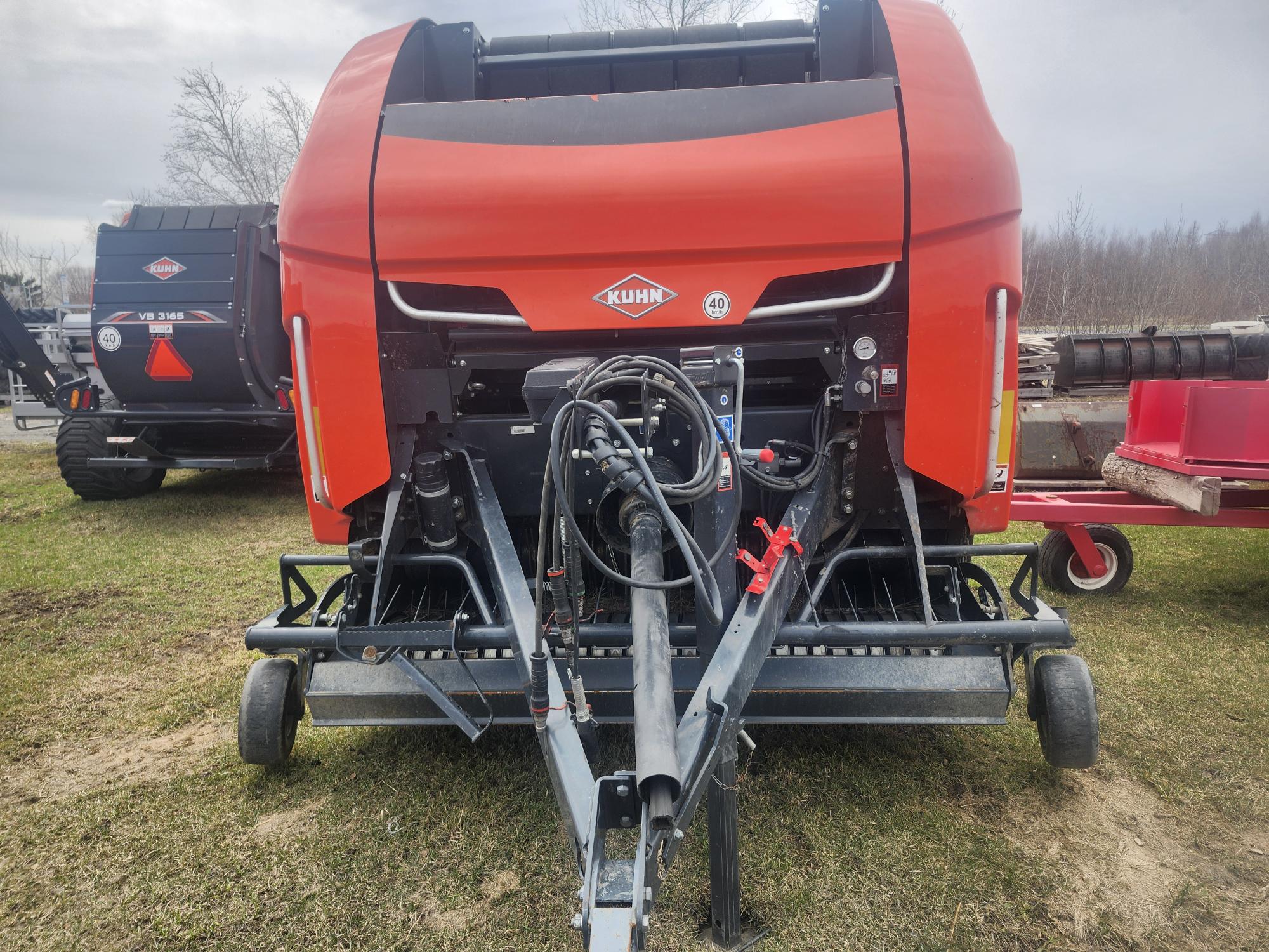 Gros plan d'une presse agricole KUHN rouge garée sur l'herbe, avec divers flexibles et connecteurs hydrauliques visibles à l'avant. D'autres équipements agricoles sont visibles en arrière-plan.