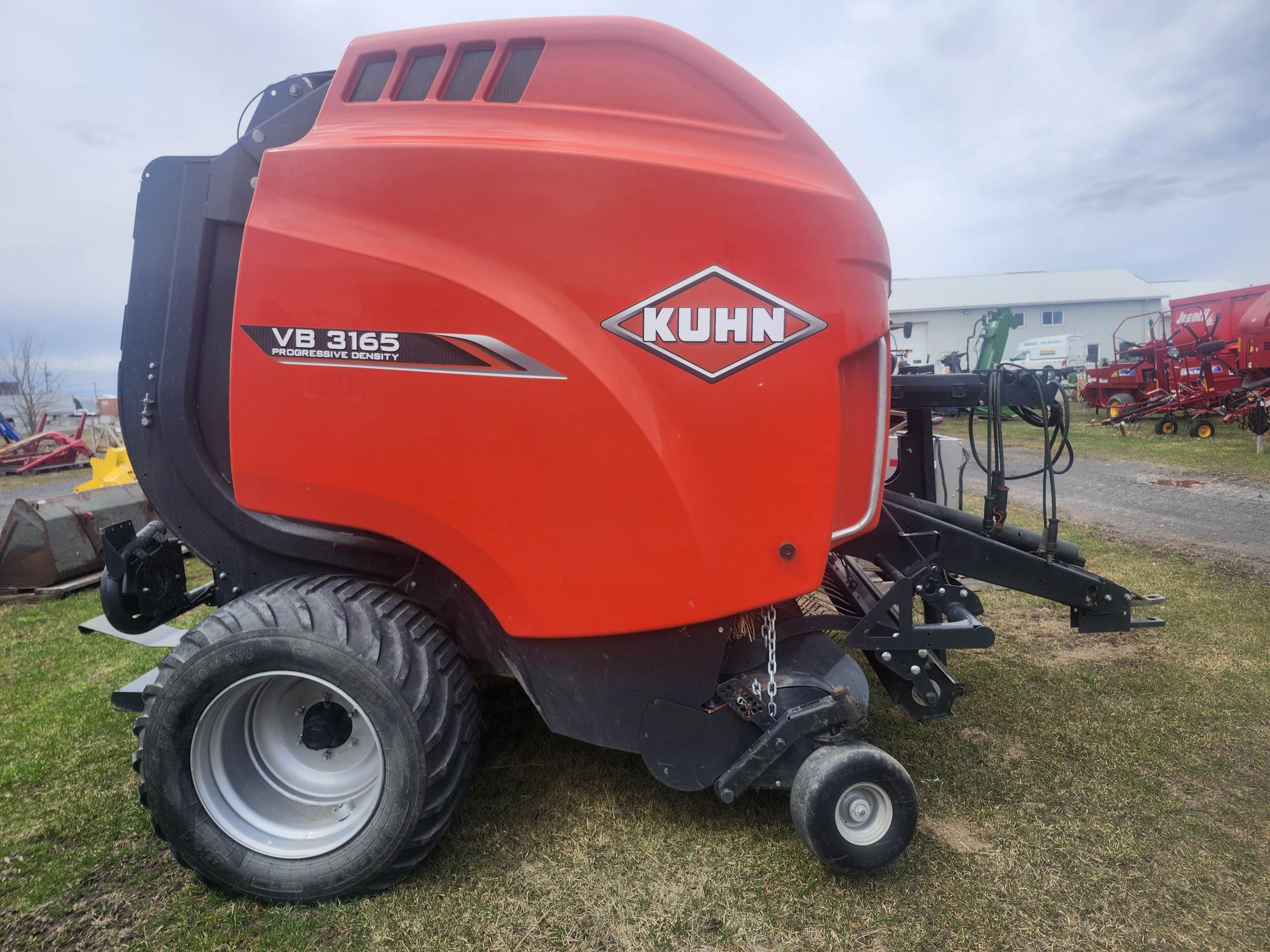 Une grande presse agricole rouge KUHN VB 3165 est posée sur l'herbe, avec de gros pneus et du matériel agricole visibles en arrière-plan sous un ciel nuageux.