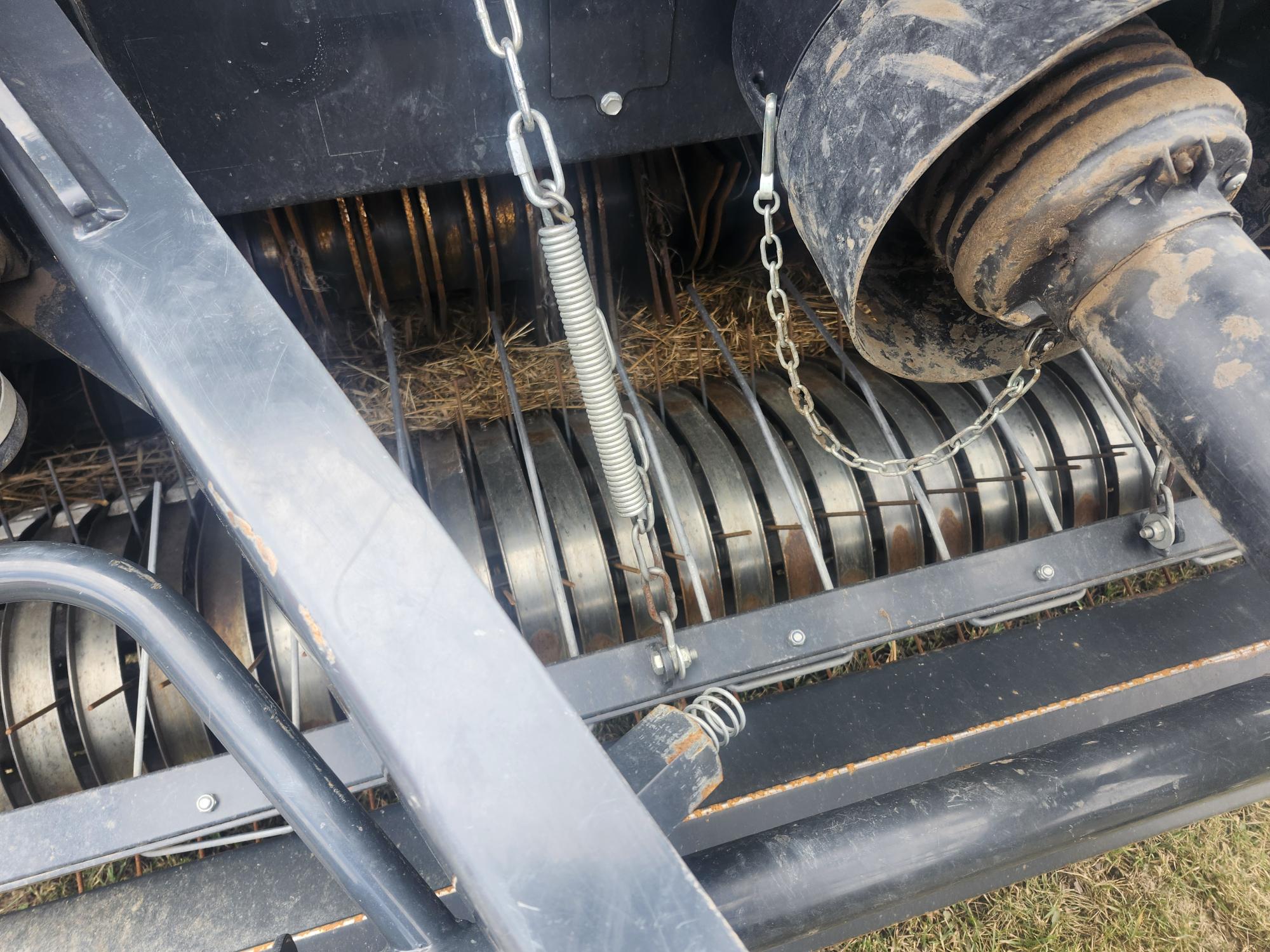 Gros plan sur des machines agricoles, montrant des rouleaux métalliques coincés entre eux par de la paille, un grand ressort métallique et des chaînes attachées à l'équipement. Les pièces semblent poussiéreuses et usées.