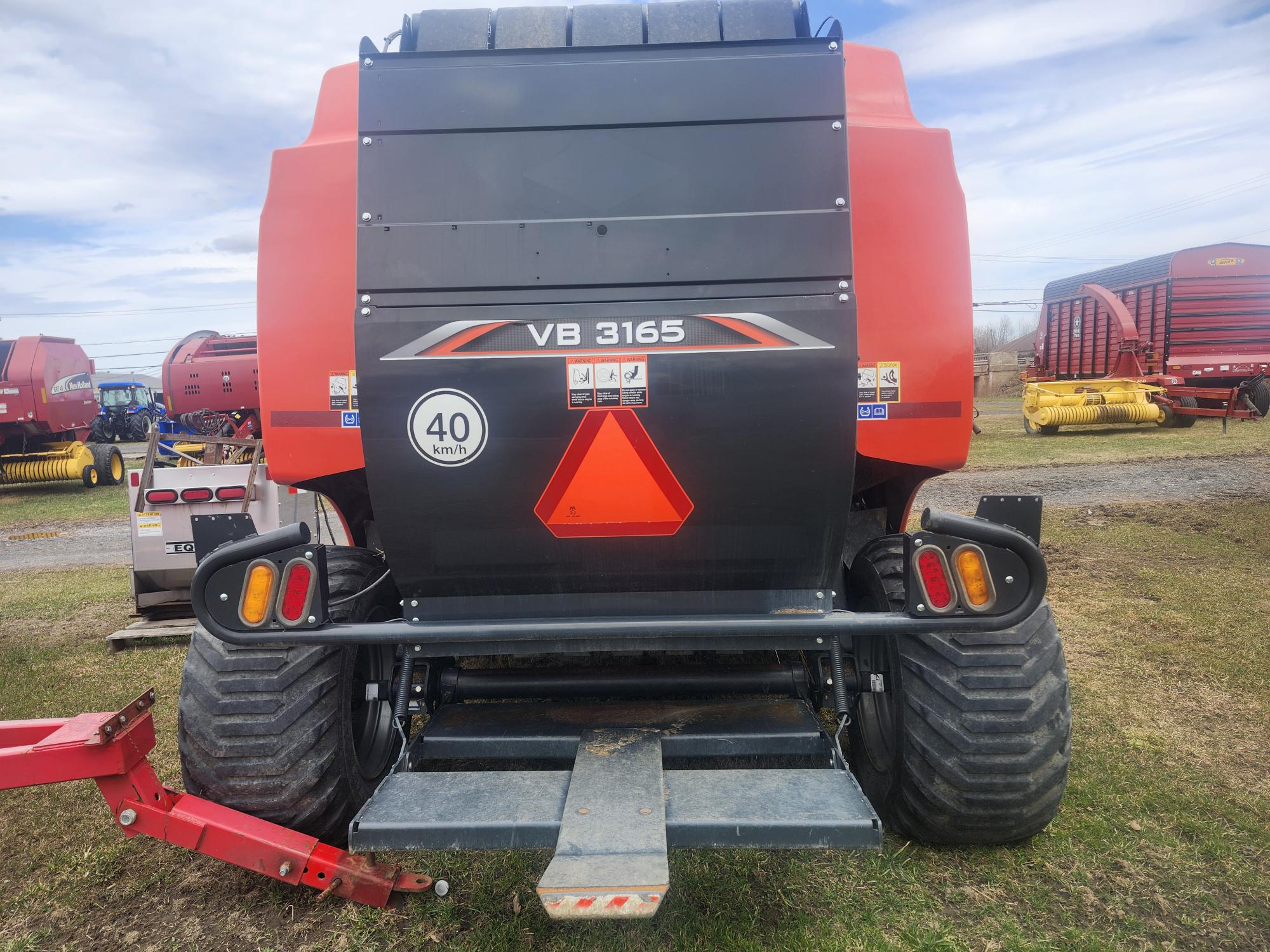 Vue arrière d'une grande presse agricole rouge et noire étiquetée « VB 3165 » avec un autocollant de vitesse de 40 km/h et un triangle orange vif pour véhicule lent, garée sur l'herbe avec d'autres machines en arrière-plan.