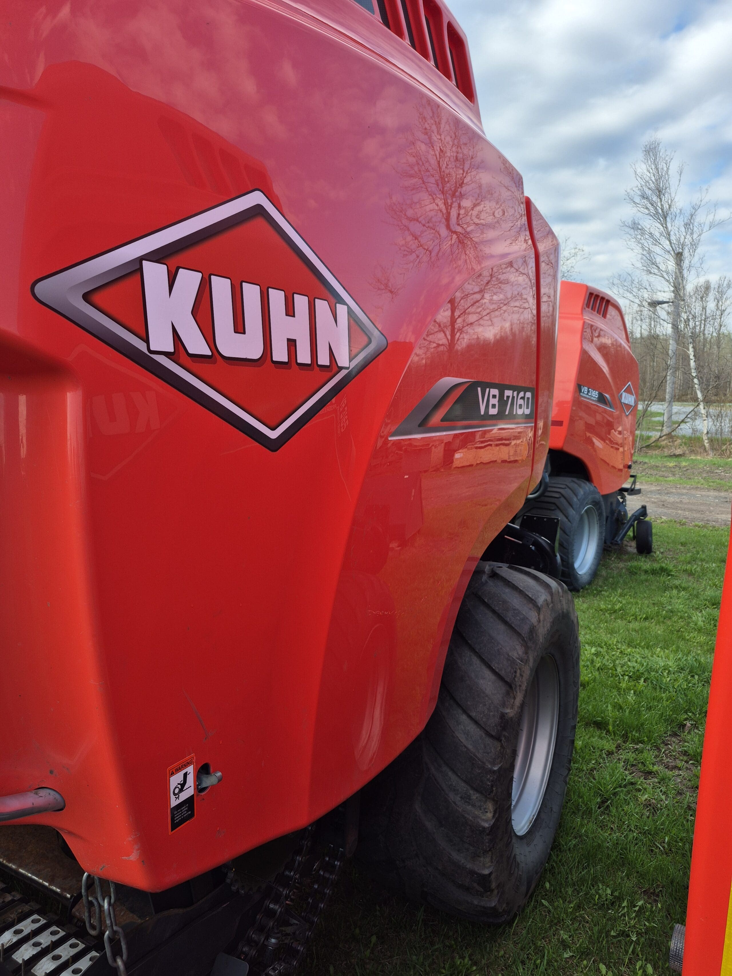 Gros plan de deux grandes machines agricoles rouges KUHN VB 7160 garées sur l'herbe, avec une machine bien en vue au premier plan et une autre à l'arrière-plan sous un ciel partiellement nuageux.