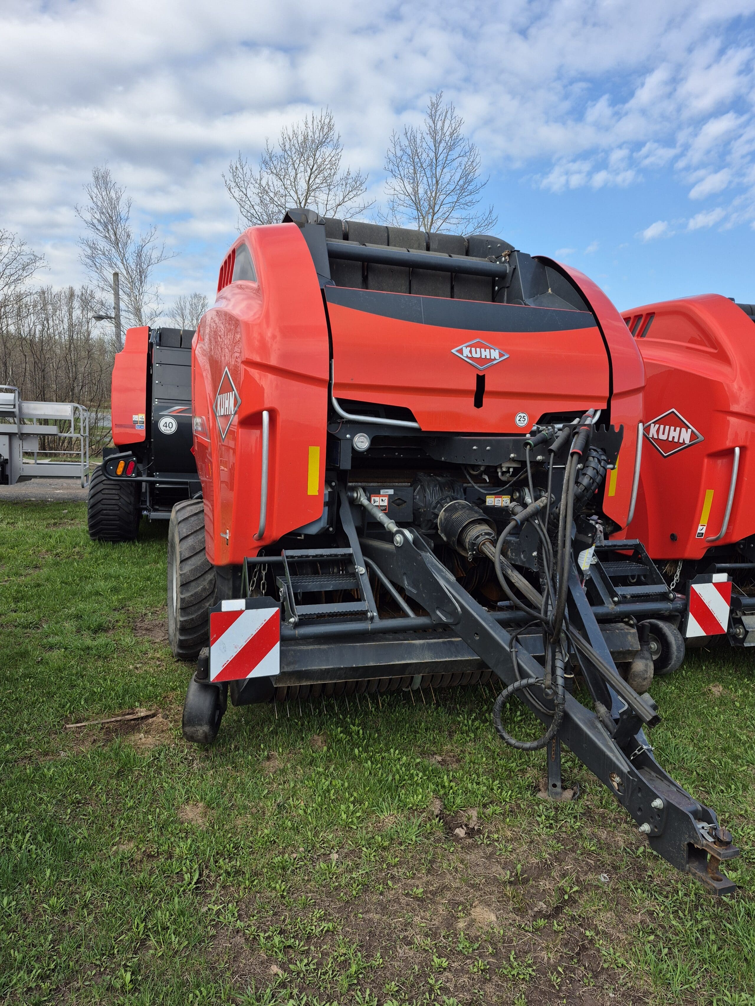 Une presse à balles rondes KUHN rouge est garée sur l'herbe avec une autre machine similaire à côté, sous un ciel partiellement nuageux avec des arbres en arrière-plan.