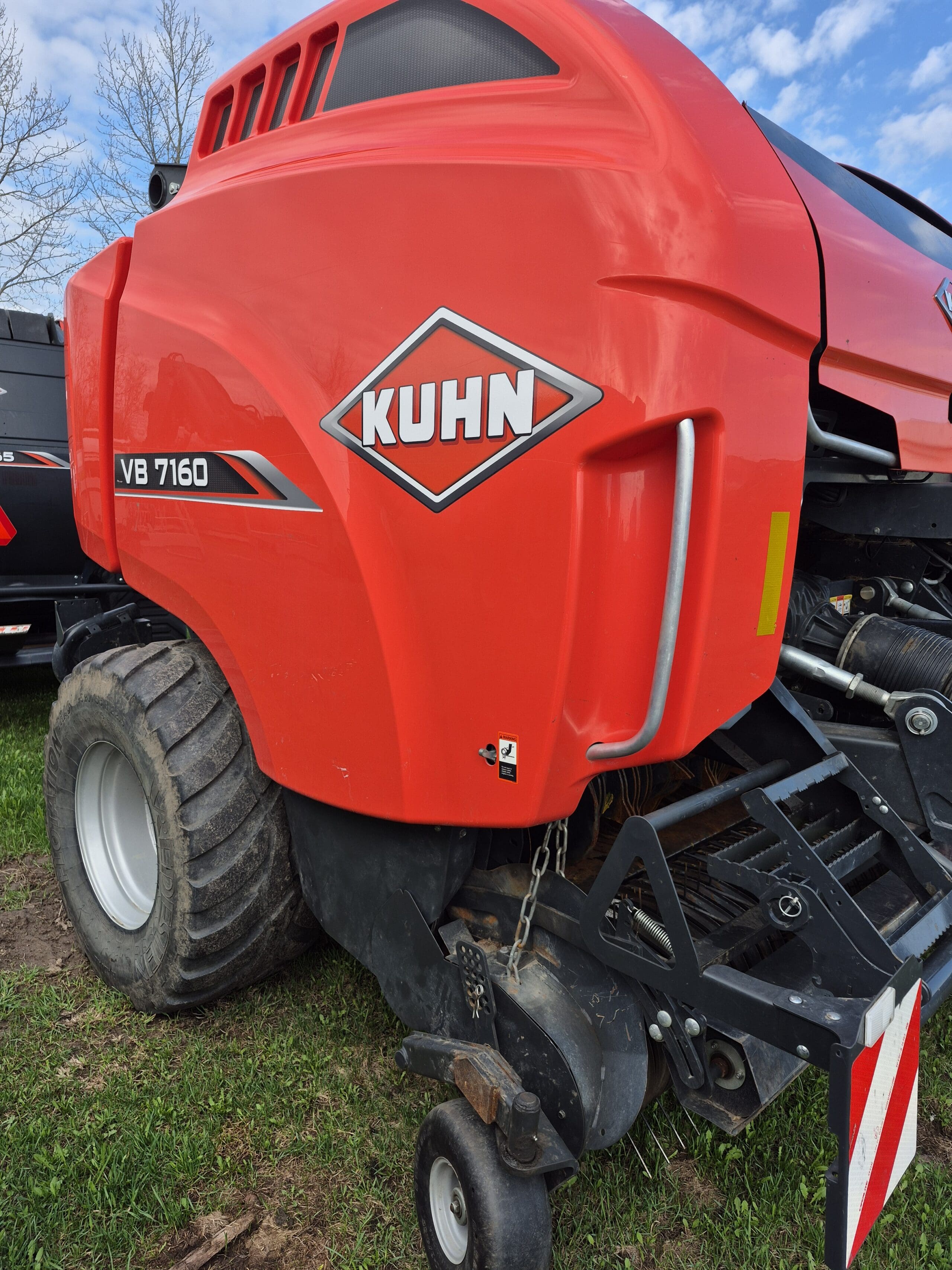 Vue rapprochée d'une grande machine agricole rouge KUHN VB 7160 avec de gros pneus, garée sur l'herbe sous un ciel partiellement nuageux.