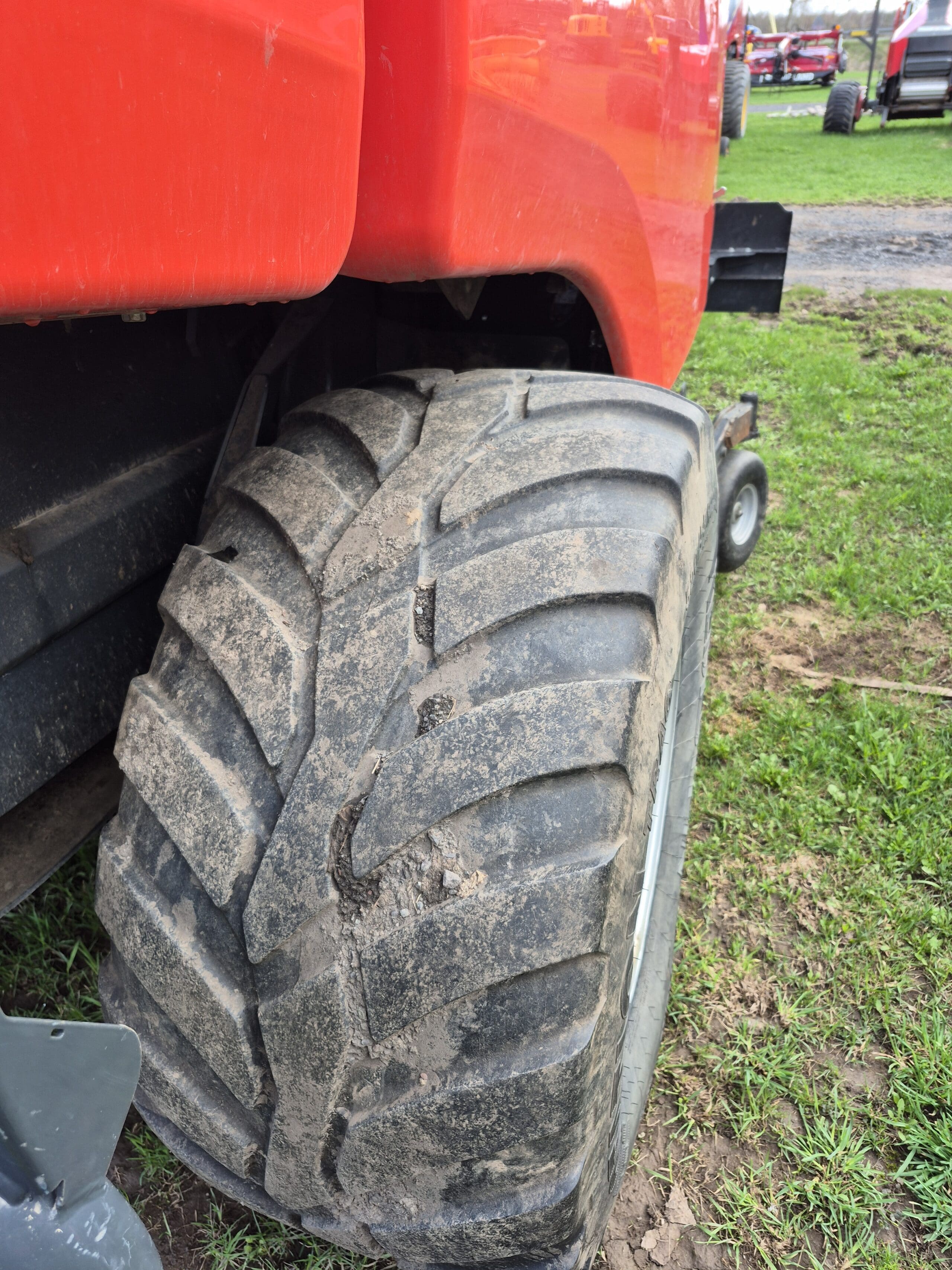 Vue rapprochée d'un gros pneu de tracteur boueux attaché à un véhicule rouge, garé sur l'herbe avec d'autres machines rouges visibles dans l'arrière-plan flou.