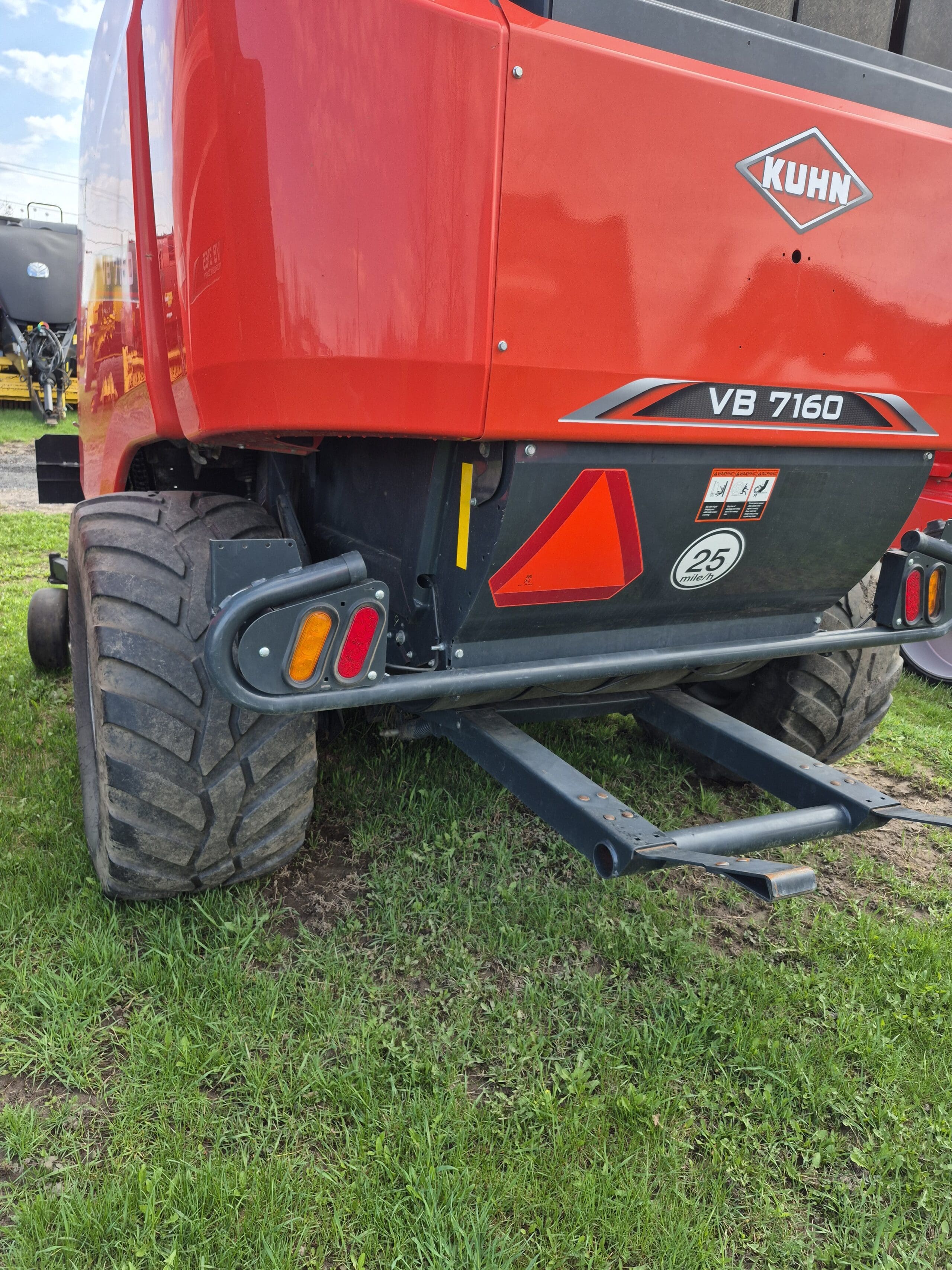 Vue arrière d'une machine agricole KUHN VB 7160 rouge avec de gros pneus, des autocollants de sécurité, un triangle de signalisation et garée sur de l'herbe verte.