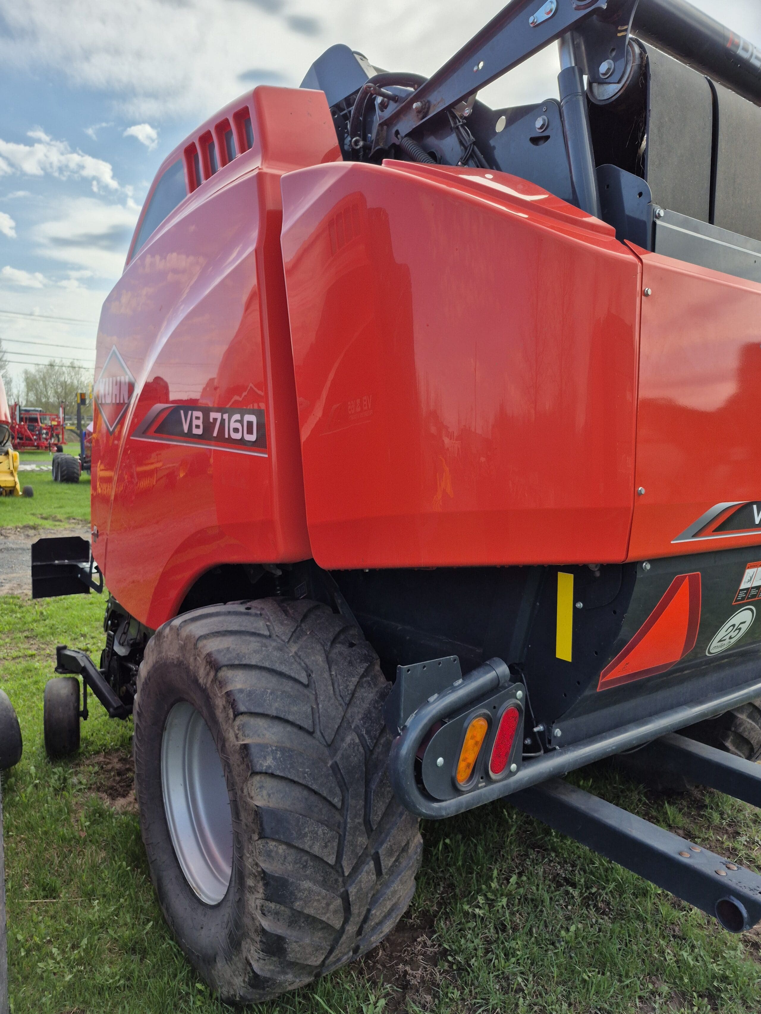 Vue rapprochée d'une machine agricole rouge, en particulier une presse à balles rondes Kuhn VB 7160, montrant sa grande roue arrière et son panneau latéral, garée sur de l'herbe verte avec un ciel nuageux en arrière-plan.
