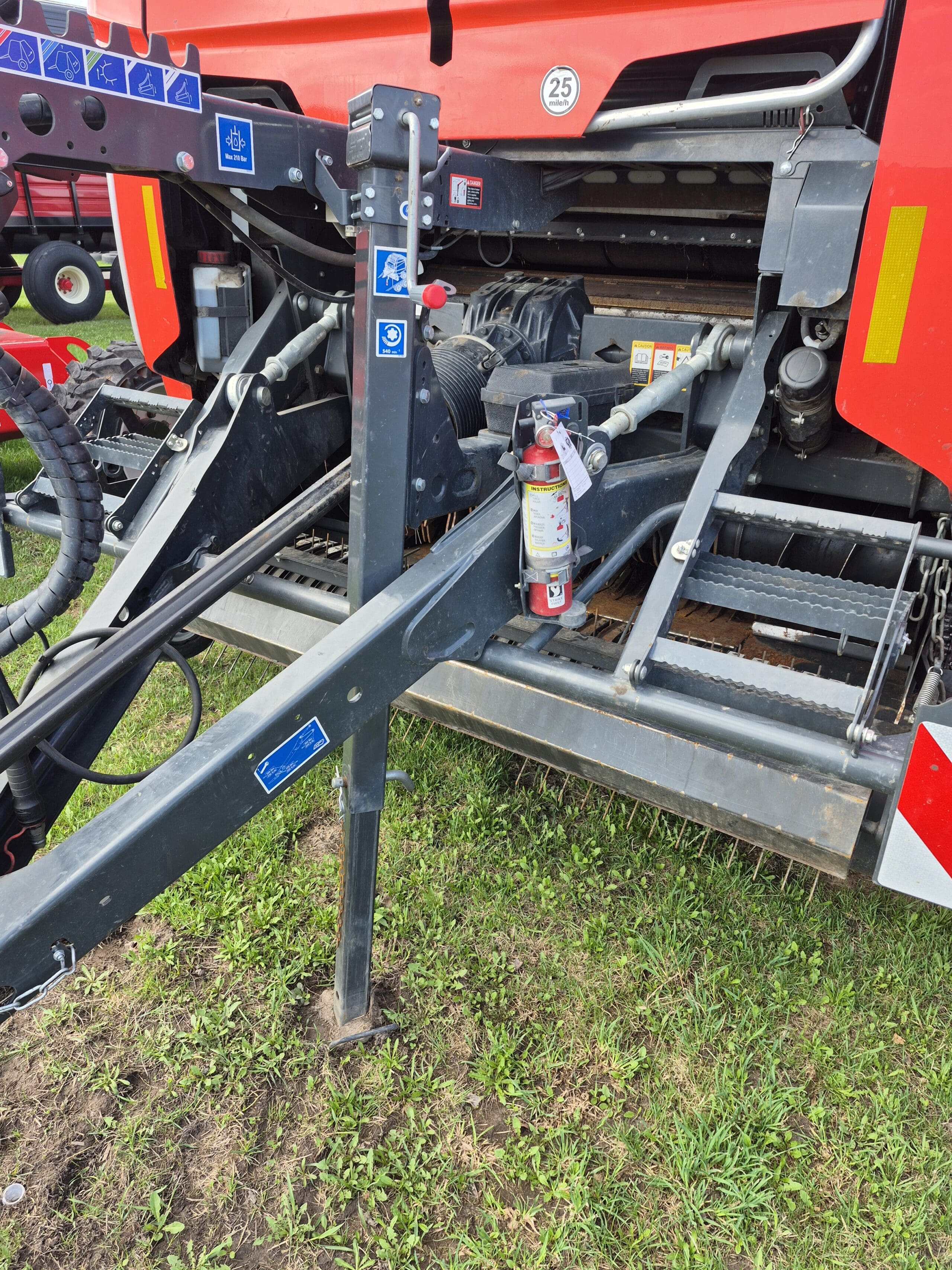 Gros plan d'une machine agricole sur de l'herbe verte, montrant un cadre métallique, des tuyaux, divers autocollants d'avertissement et d'instructions, un extincteur monté sur le cadre et des pièces d'une machine agricole rouge en arrière-plan.
