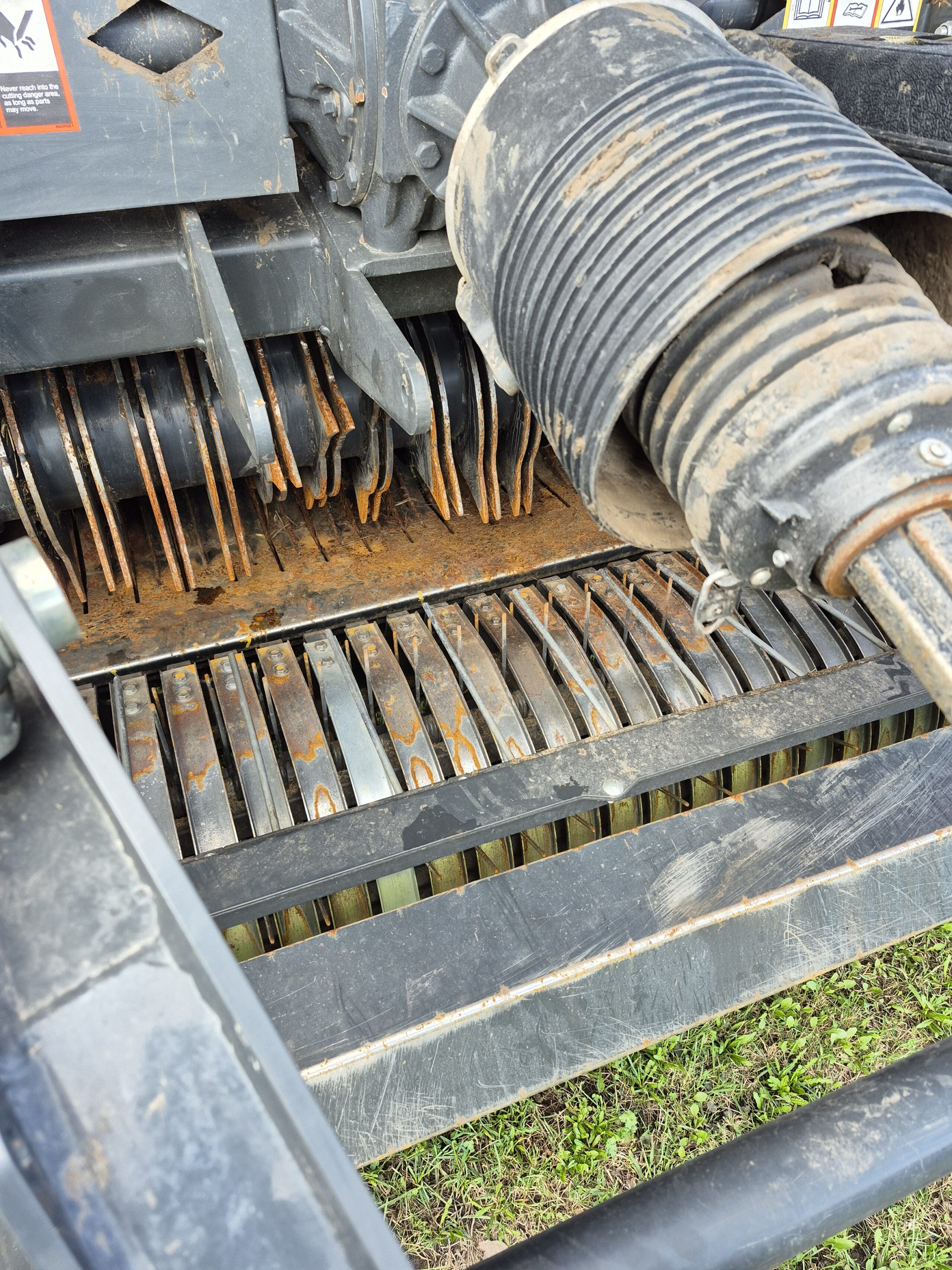 Gros plan d'une machine agricole montrant des lames ou des dents métalliques, un arbre rotatif, ainsi que de la rouille et de la saleté visibles. L'équipement est positionné au-dessus de l'herbe verte.