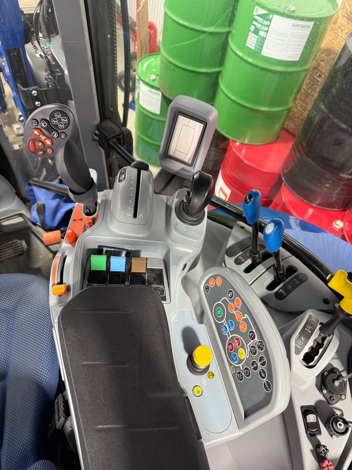Cockpit d'un tracteur moderne avec diverses commandes, dont des leviers, des boutons et un écran d'affichage. Des boutons multicolores et un joystick sont visibles. Des barils verts sont empilés en arrière-plan.