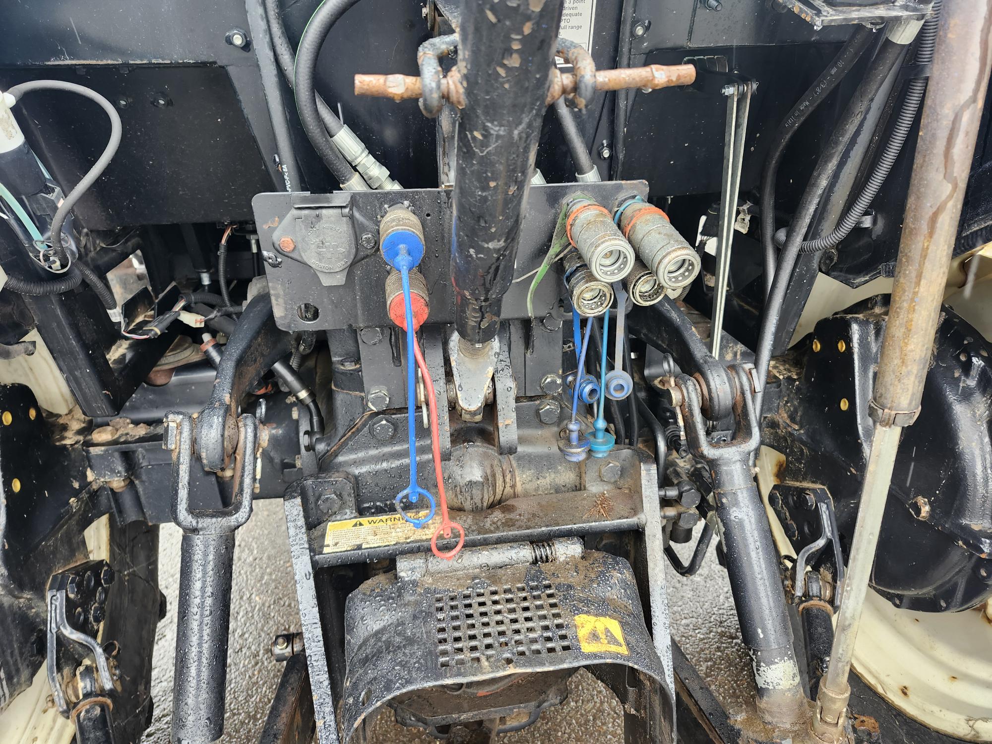 Gros plan sur le système hydraulique et l'attelage du tracteur, montrant divers flexibles, leviers et connecteurs. L'équipement est noir et métallique, avec des conduites et des vannes de couleur rouge et bleue, indiquant les composants permettant de fixer le matériel agricole.