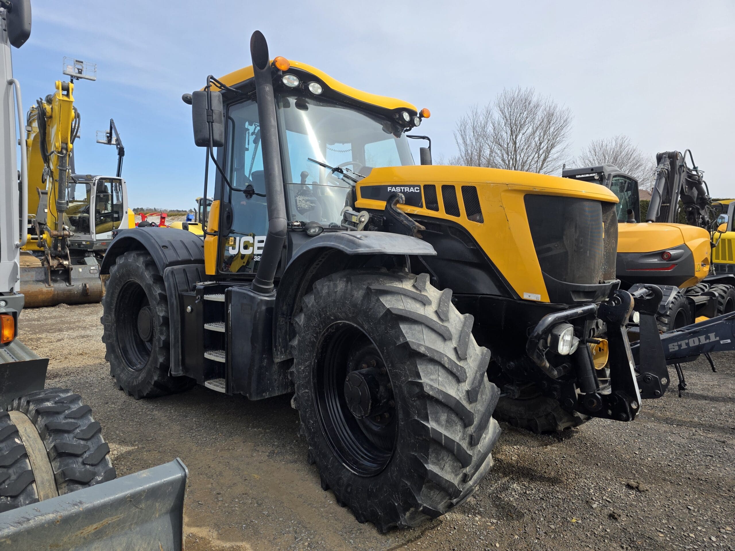 Un gros tracteur JCB Fastrac jaune et noir avec des pneus surdimensionnés est garé sur un terrain en gravier, entouré d'autres véhicules et équipements de construction.