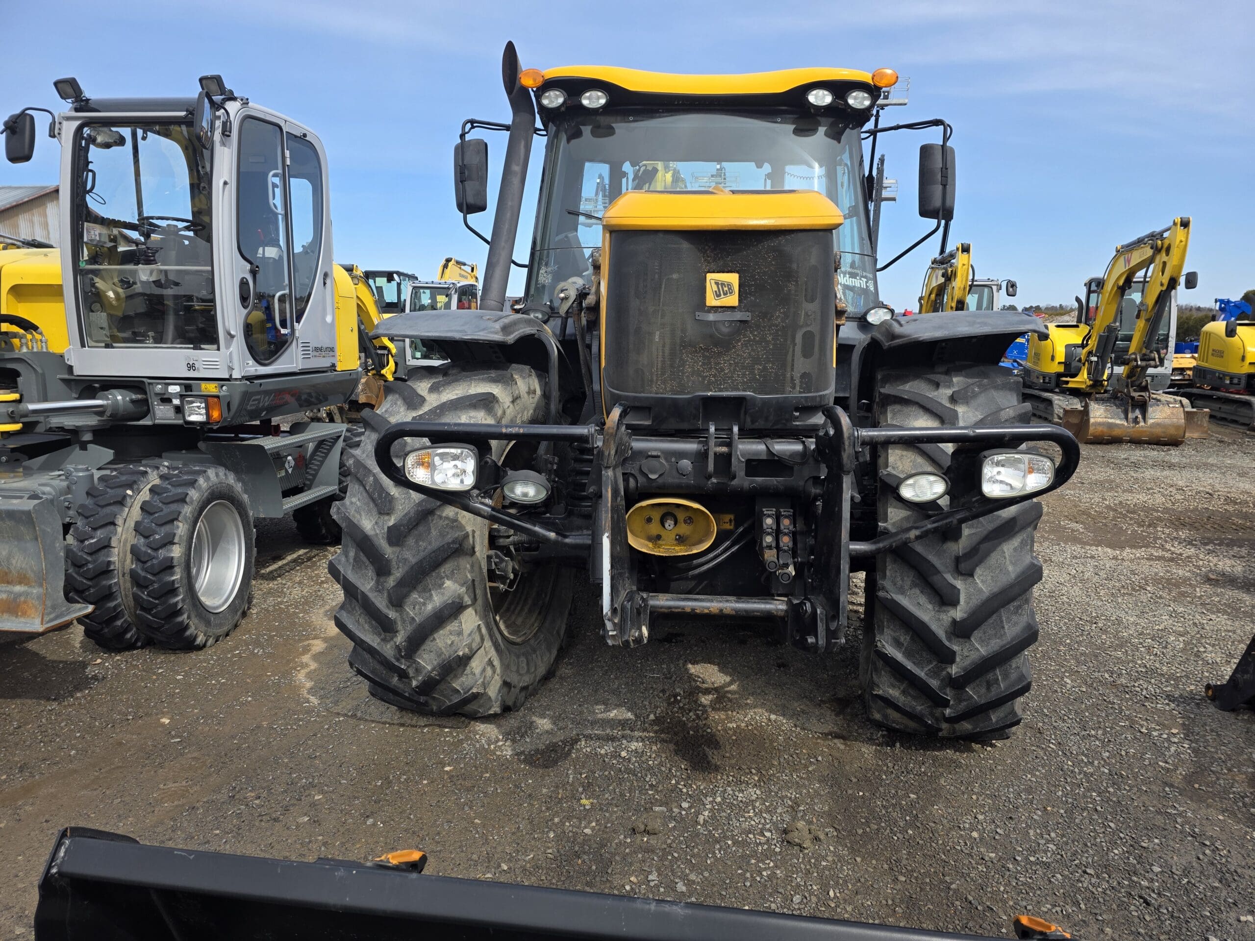 Un gros tracteur jaune et noir est garé sur un terrain en gravier, avec d'autres véhicules et équipements de construction visibles en arrière-plan sous un ciel clair.