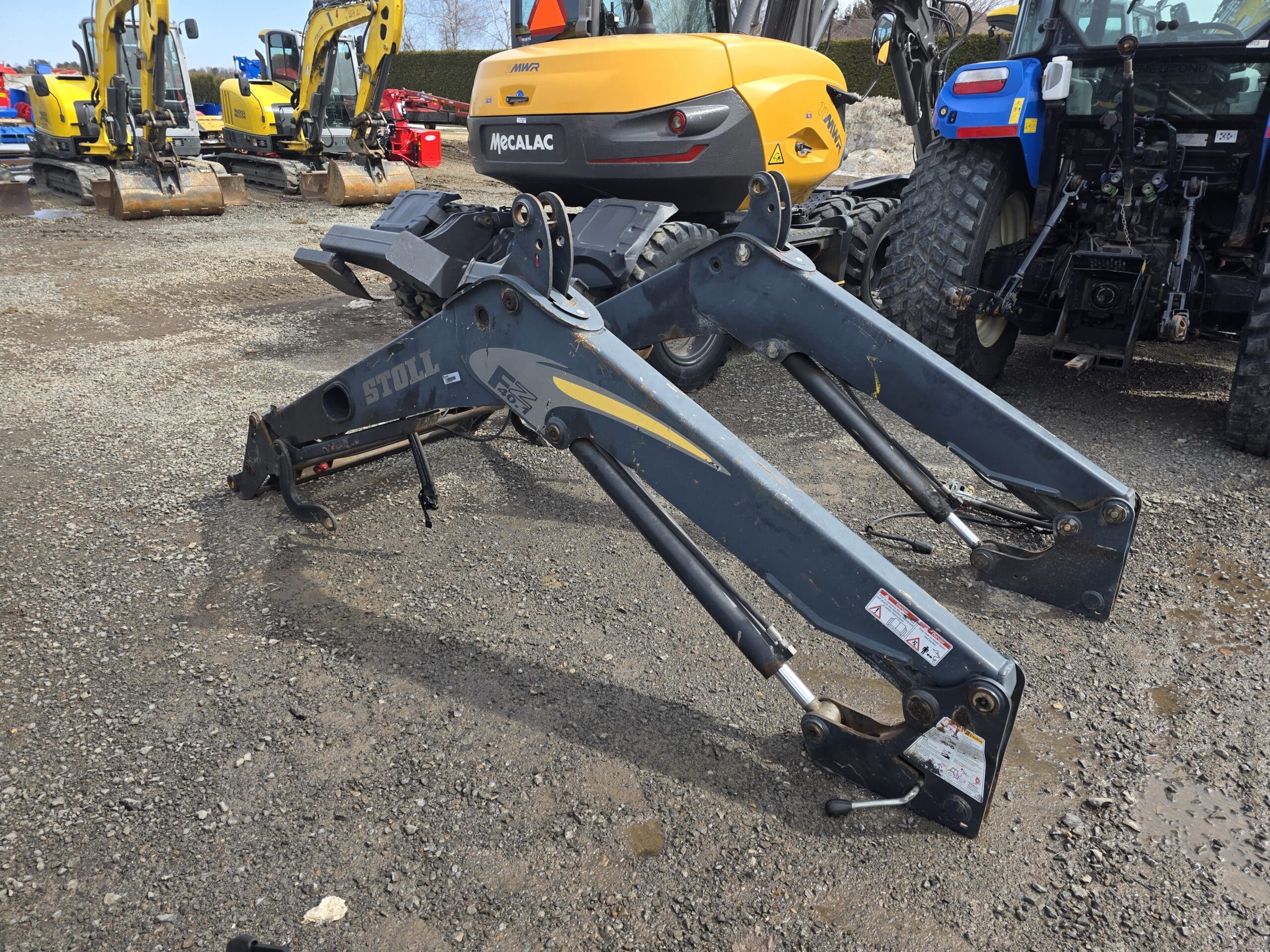 Deux grands bras de chargement métalliques détachés avec vérins hydrauliques reposent sur du gravier dans une cour entourée d'équipements de construction, notamment des excavatrices jaunes et un tracteur bleu.