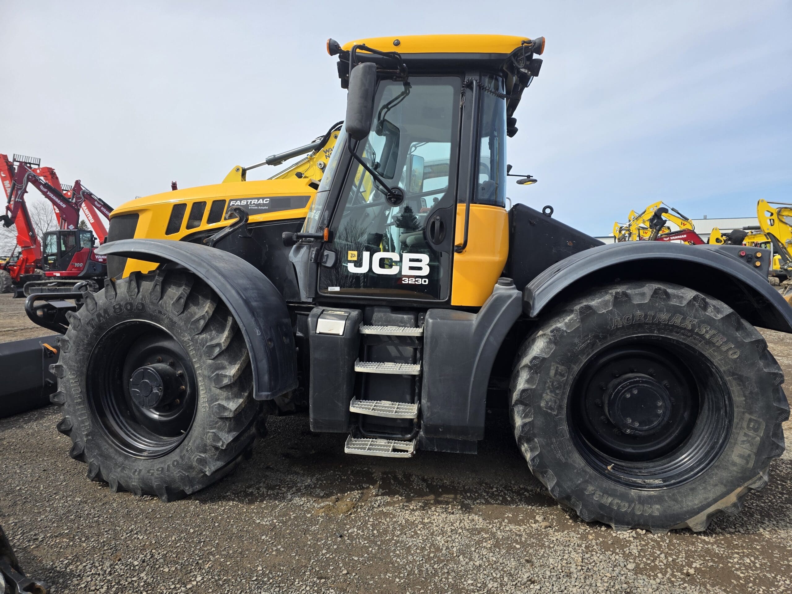 Un gros tracteur JCB Fastrac 3230 jaune et noir avec de gros pneus est garé sur du gravier, entouré d'autres véhicules et machines de construction en arrière-plan.